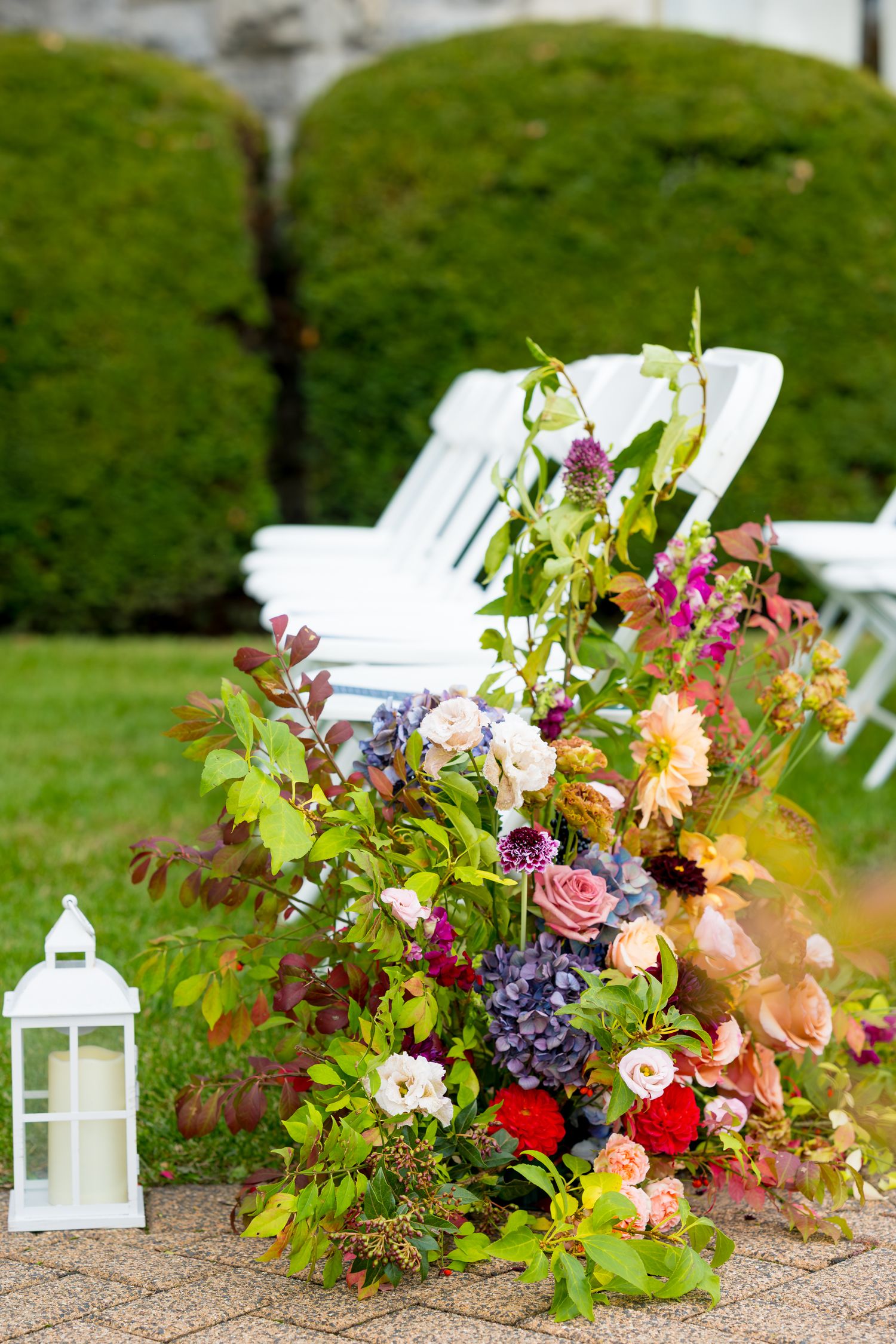 Colorful floral arrangements with gerbera daisies and mixed blooms line a garden path next to white chairs and lanterns.
