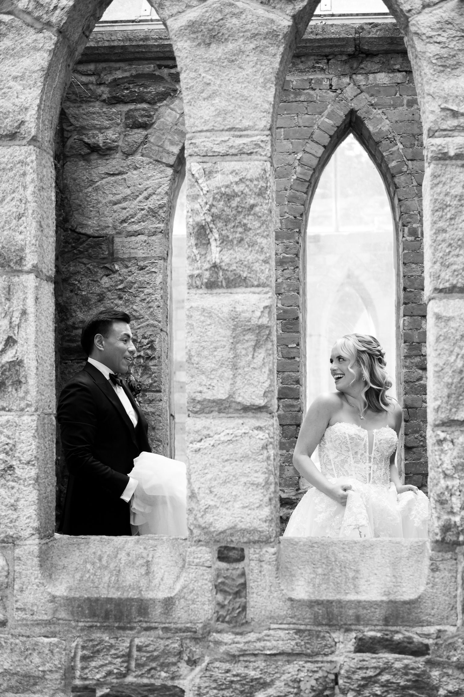 A bride and groom pose in separate stone church window arches in an artistic black and white wedding photograph.