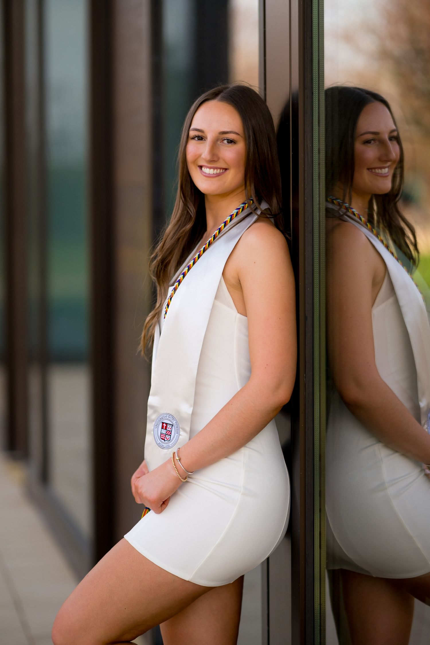 Sacred Heart University graduate in white dress poses against modern building for graduation photos.