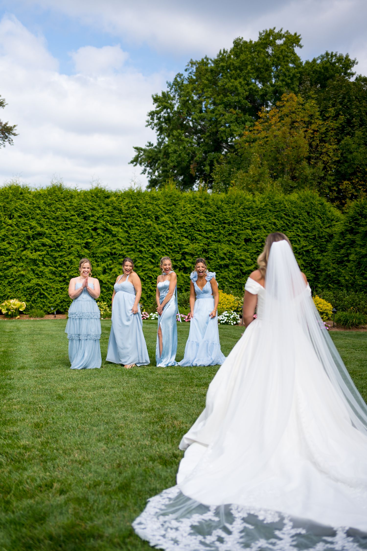 Wedding party in light blue bridesmaid dresses pose on a lush green lawn with flowing white bridal gown train.