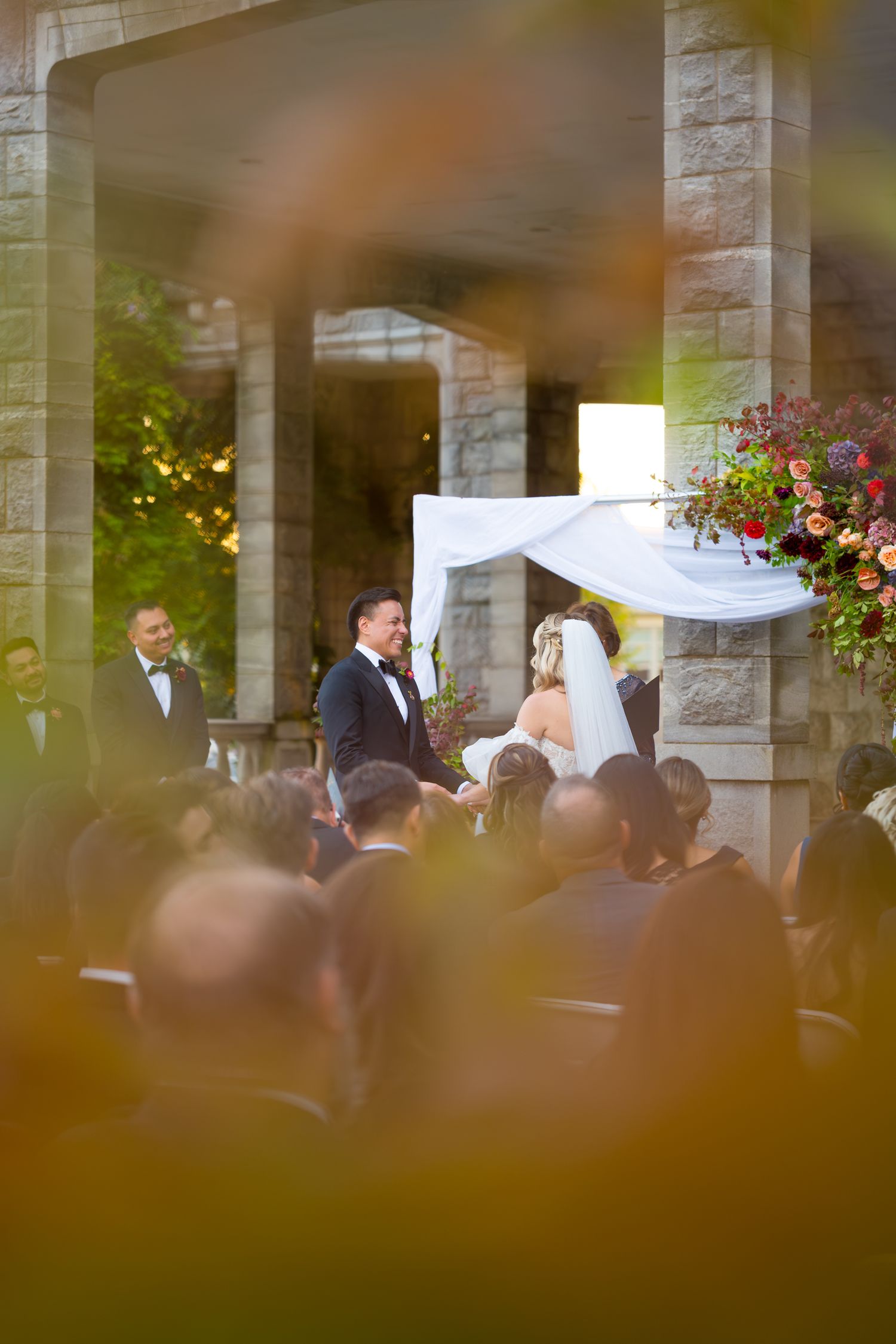 Wedding ceremony taking place outdoors under a stone colonnade with floral decorations and guests in attendance.
