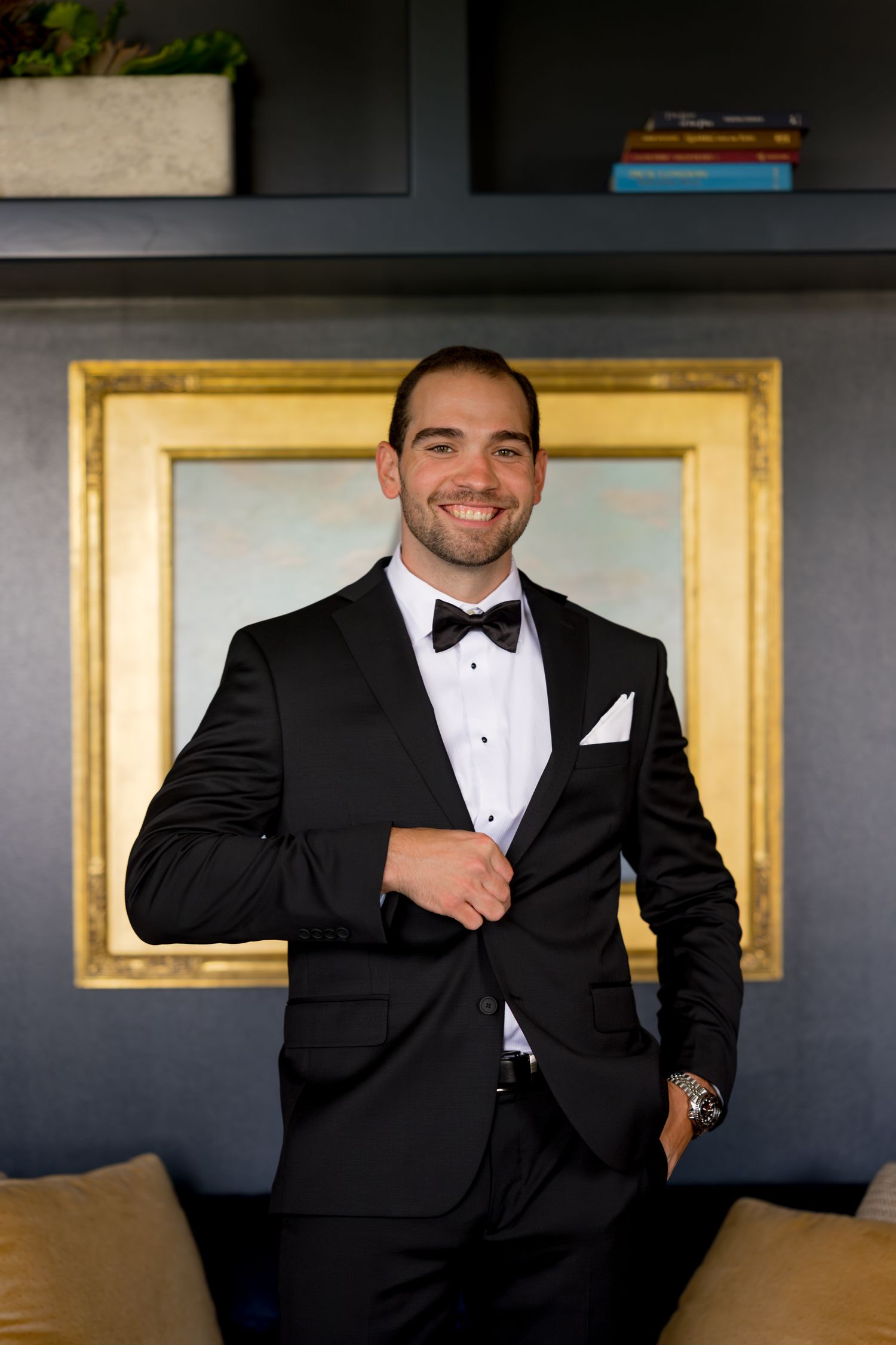 Groom in black tuxedo poses against dark wall with gold-framed artwork.