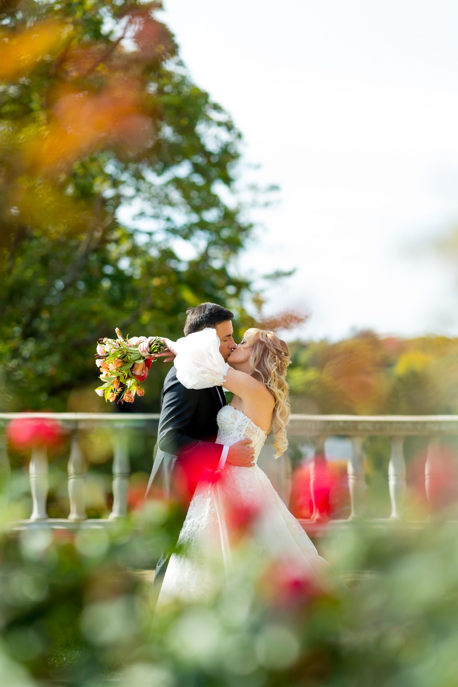 Romantic wedding portrait through red flowers with couple embracing in garden setting
