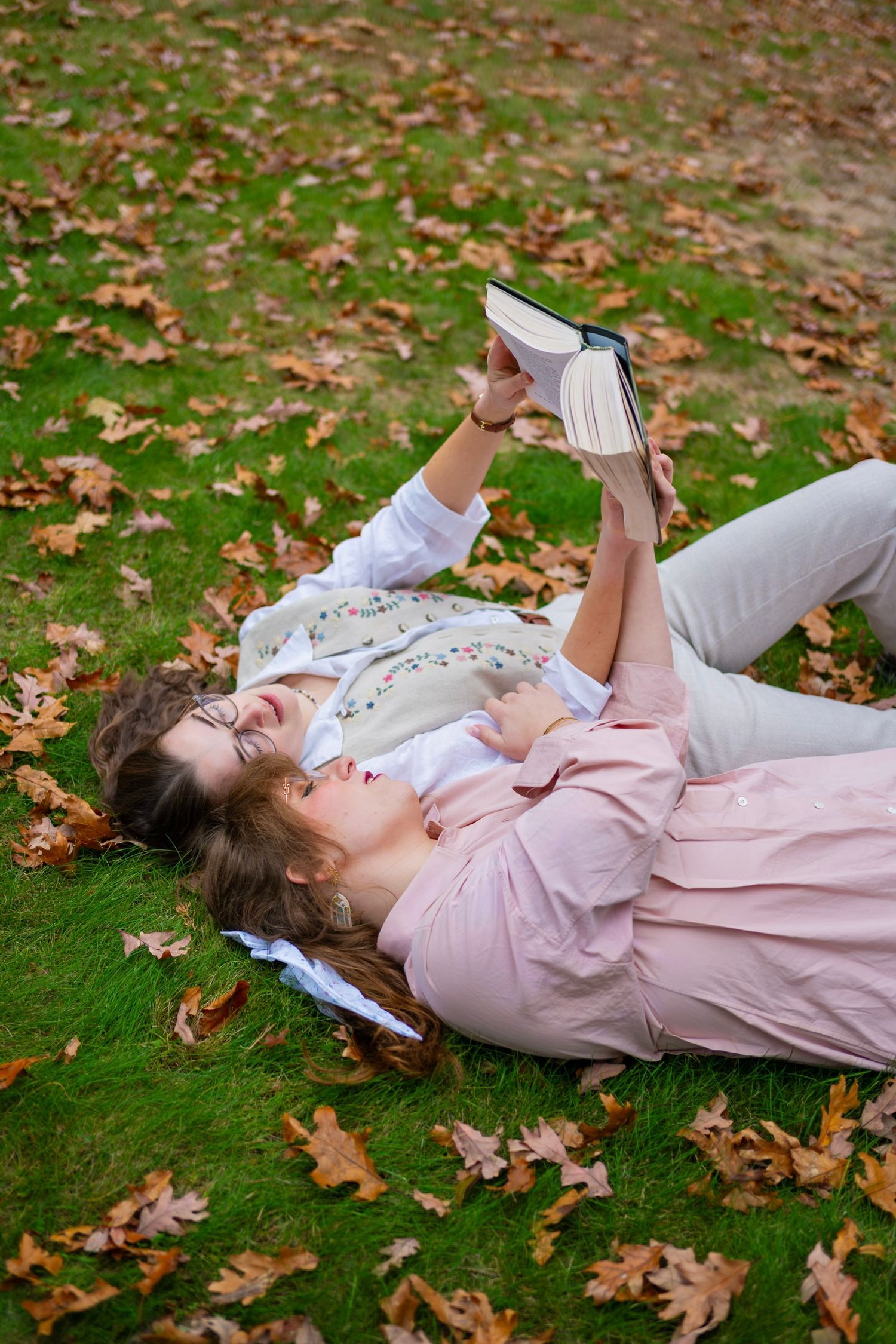 Couple lying on autumn leaves reading a book in a relaxed outdoor setting.