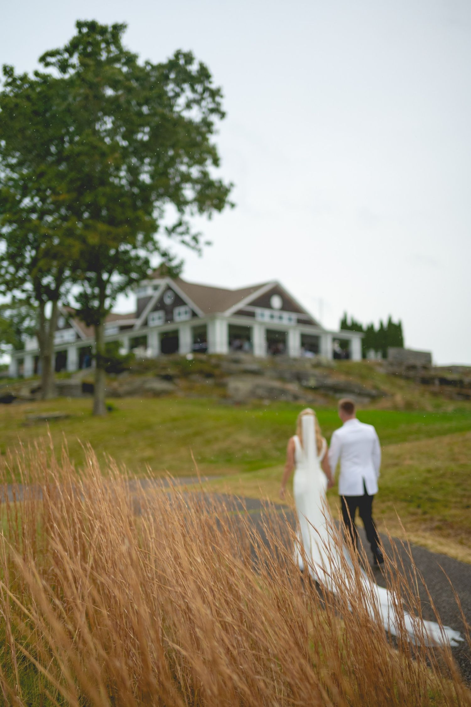 Silhouettes of a wedding couple walking through tall grass with a house on a hill in the background.