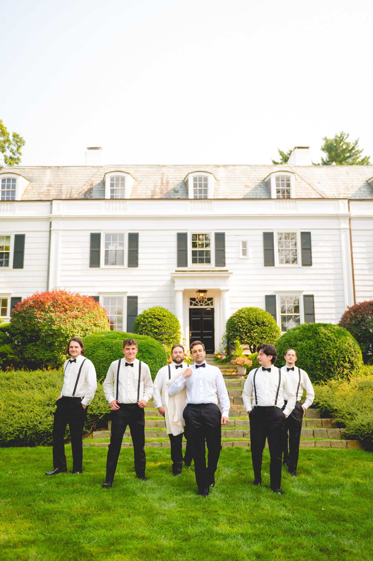 Groomsmen in classic black and white attire lined up on manicured lawn of elegant venue.