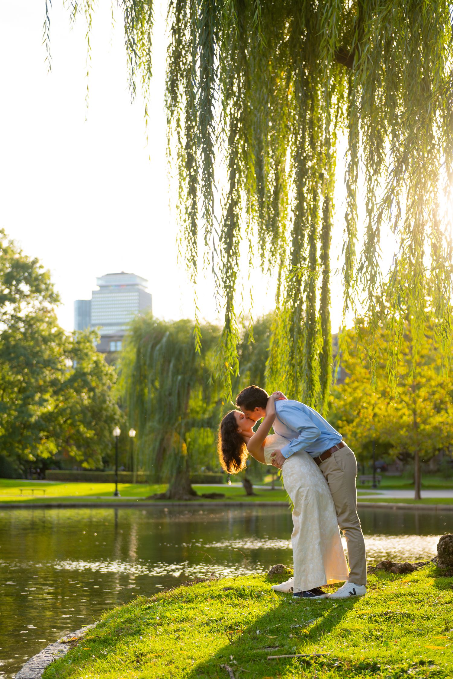 A romantic couple embraces under a weeping willow tree by a pond in a park during golden hour.
