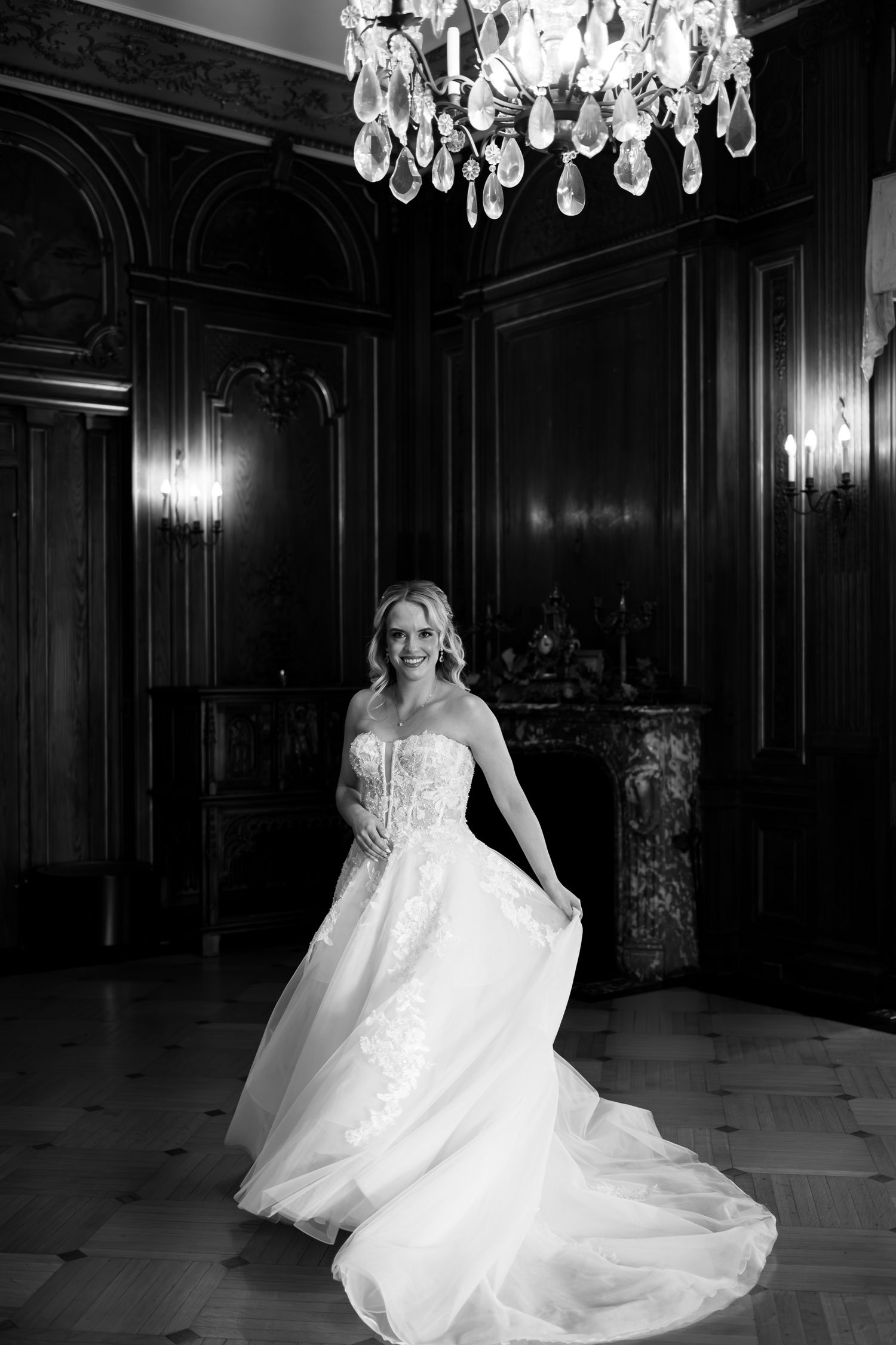 A bride in a flowing white wedding gown poses elegantly under a crystal chandelier in a dark, ornate ballroom.