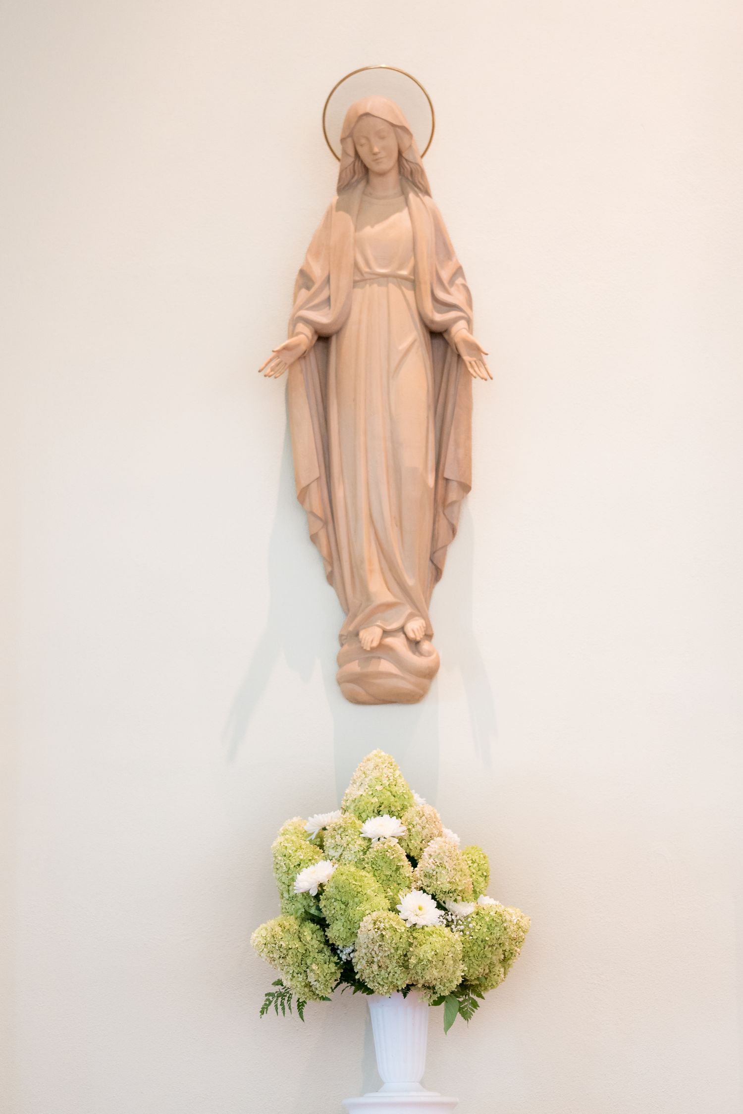 A pale religious statue mounted on a wall above a vase of cream and green hydrangea flowers.