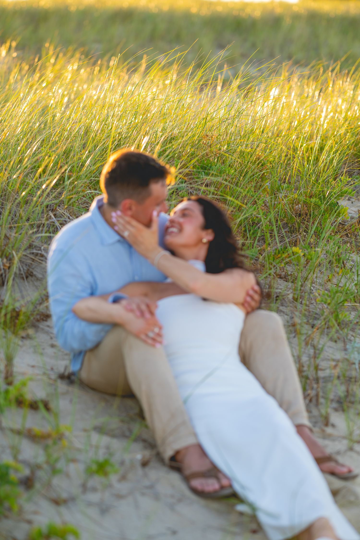 Couple embracing and kissing on sandy beach with grass during sunset.