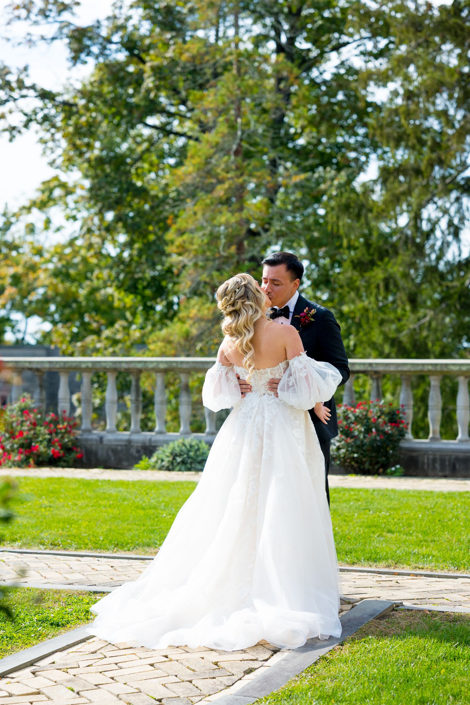 Couple embraces during outdoor wedding ceremony on stone patio with white railing and garden backdrop.