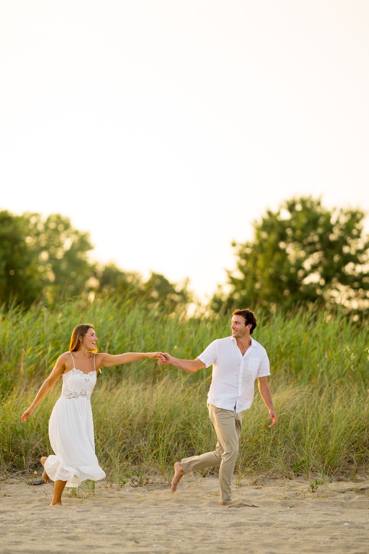 Playful couple holding hands while running on beach path through tall grass.