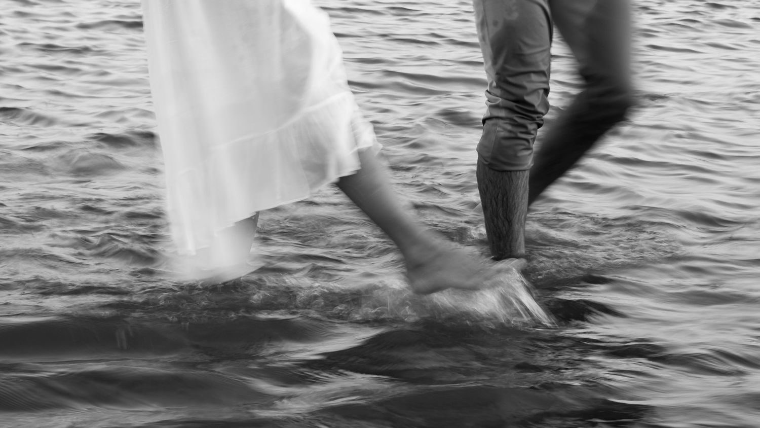 A person in a wedding dress splashes through shallow water creating ripples and movement in black and white.