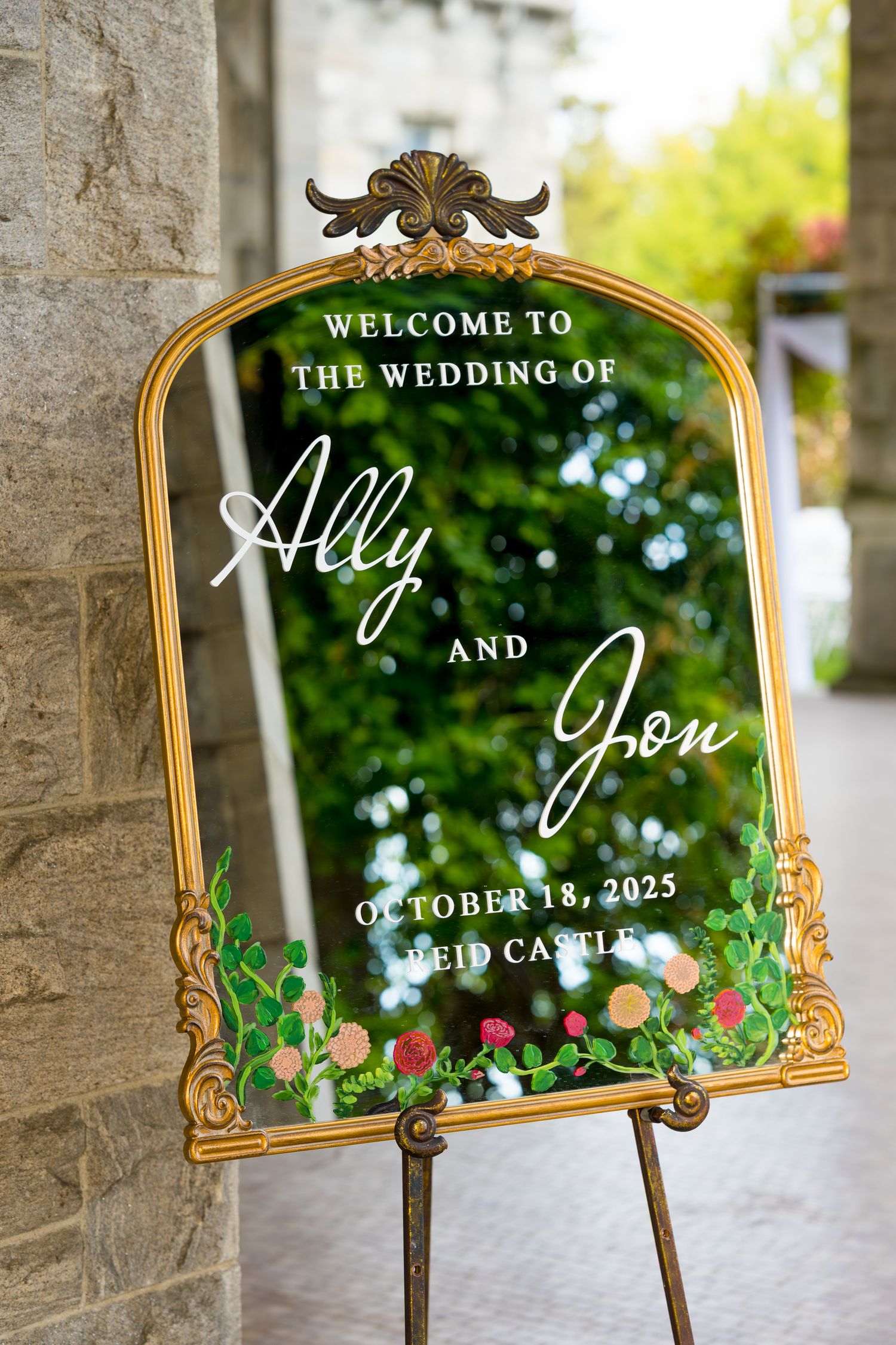 Elegant gold-framed wedding welcome signs with ornate designs and green floral accents displayed against a stone wall.