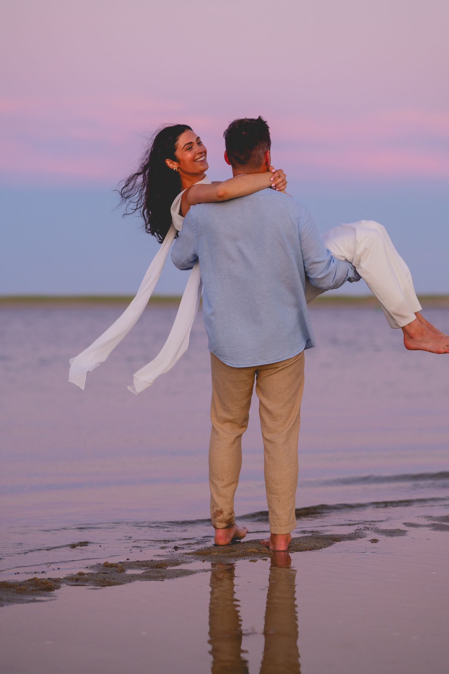 Couple dancing together on a beach at sunset with pink and purple skies creating a romantic atmosphere.