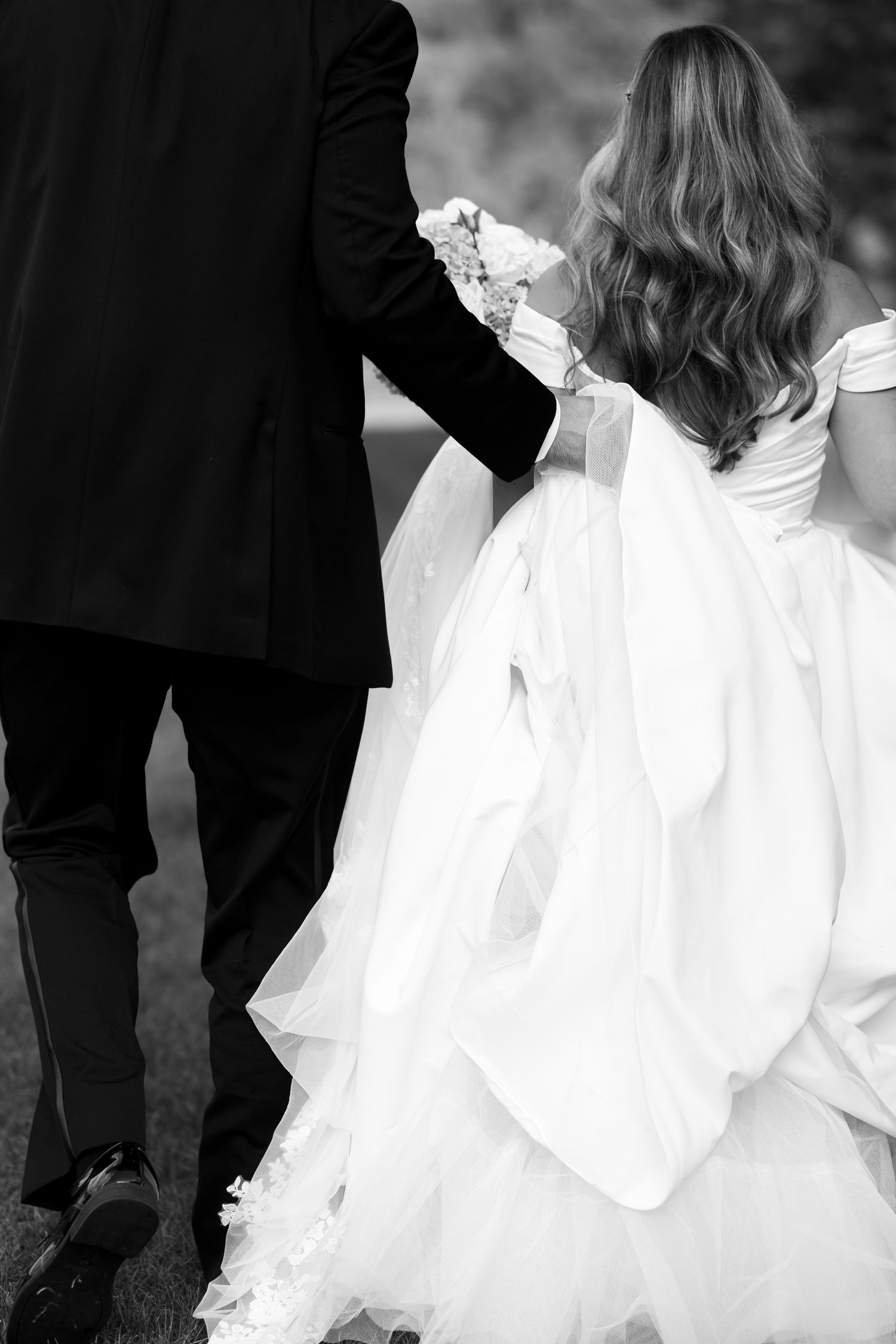 Black and white wedding photo showing couple walking with flowing white dress and formal attire visible from behind.