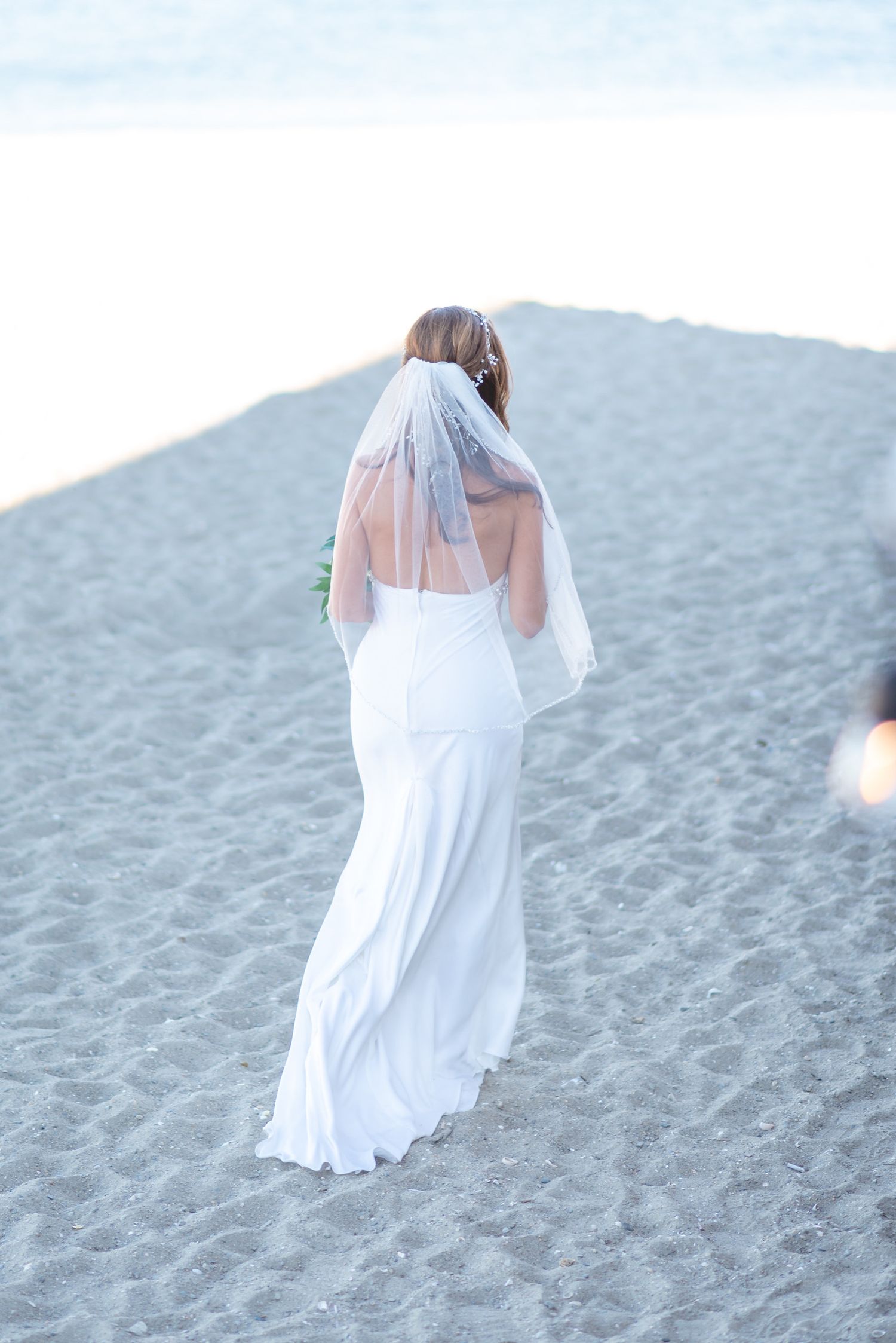 Bride in flowing white dress walks along sandy beach with veil flowing in the breeze.