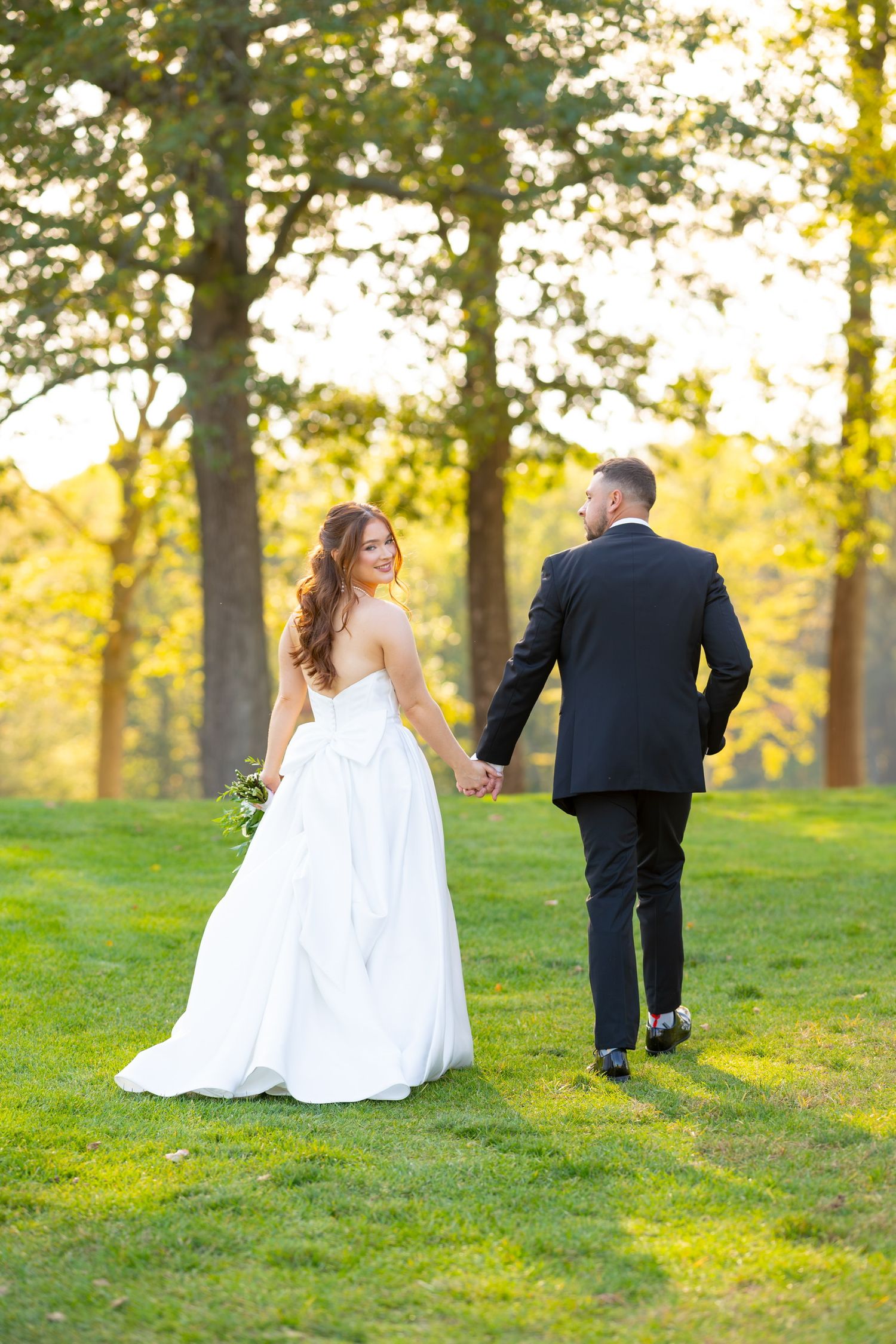 Bride and groom walk together on grassy hill with golden sunlit trees in background.