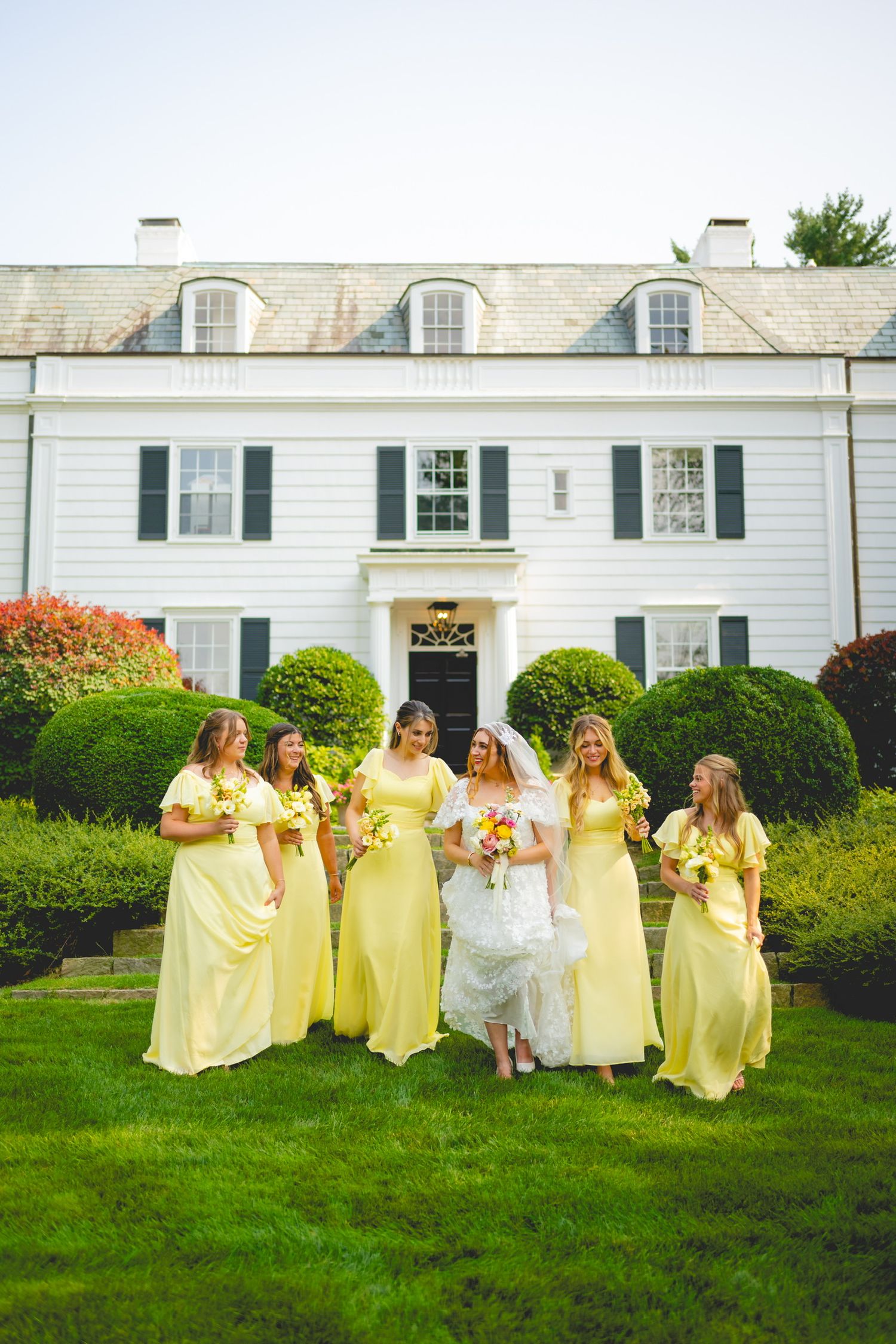 Wedding party in yellow bridesmaid dresses poses on lawn in front of white colonial building.