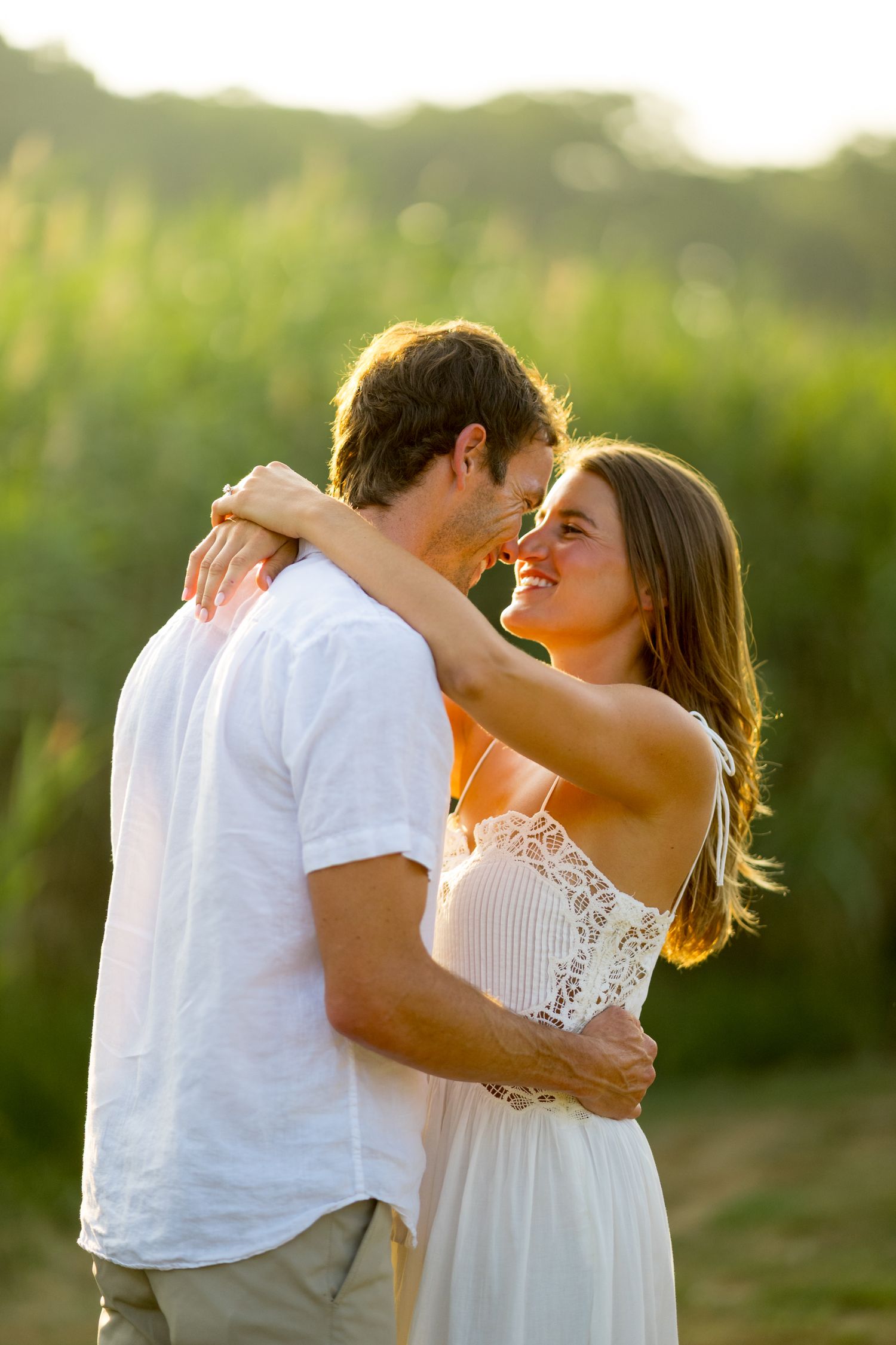 A couple in white clothing embraces in a romantic moment during golden hour in a natural outdoor setting.