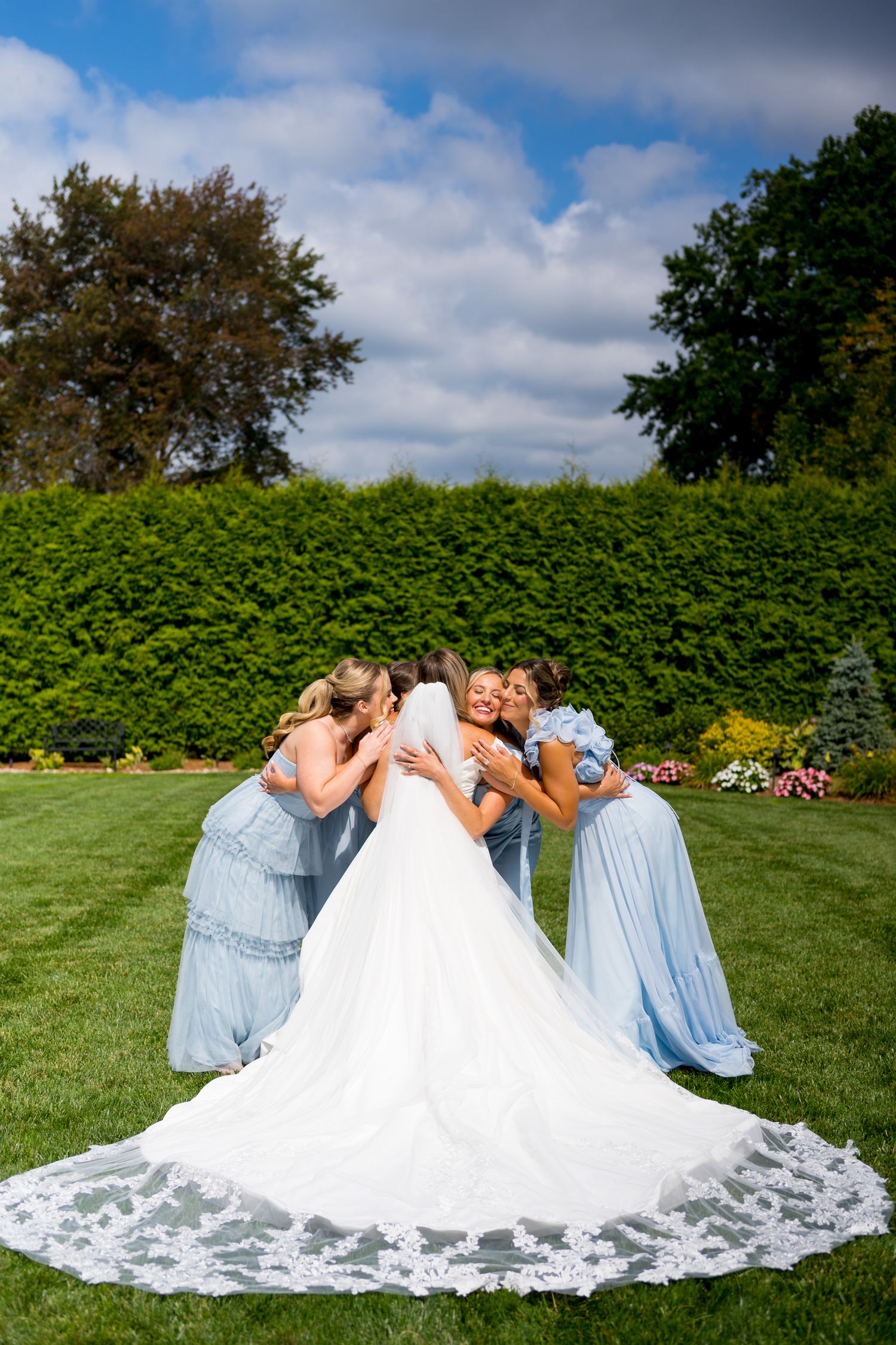 Wedding party in light blue dresses gather around bride's lace train on green lawn.