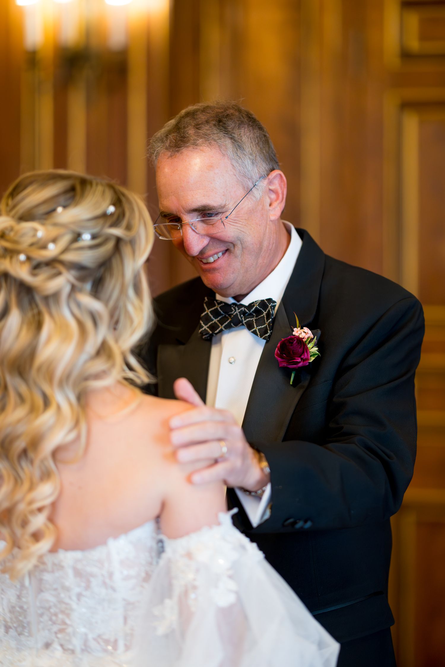 An emotional moment between a bride in a white lace dress and a gentleman in a formal suit with a red boutonniere.