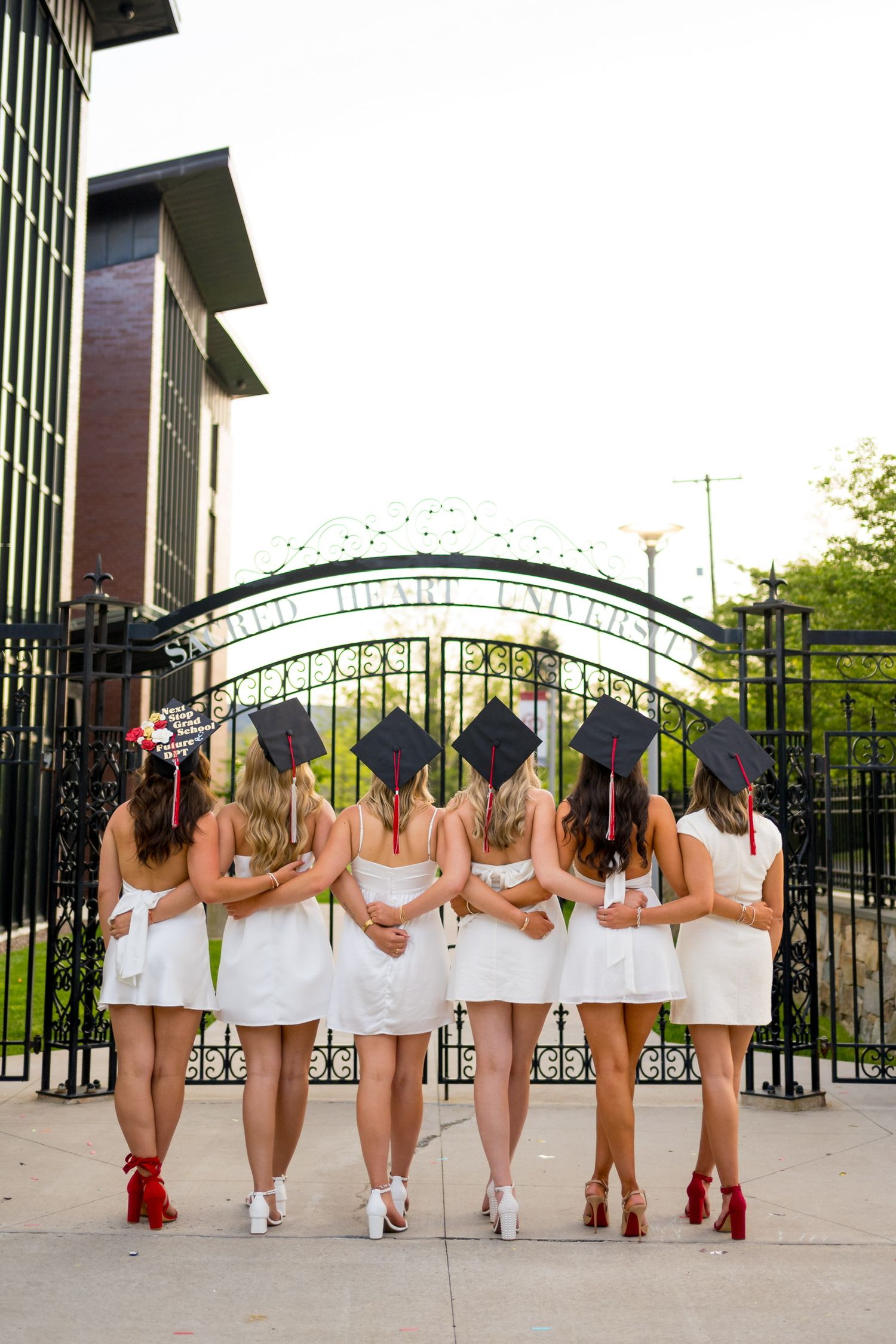 Row of women in white dresses posing with red heels against iron gate.