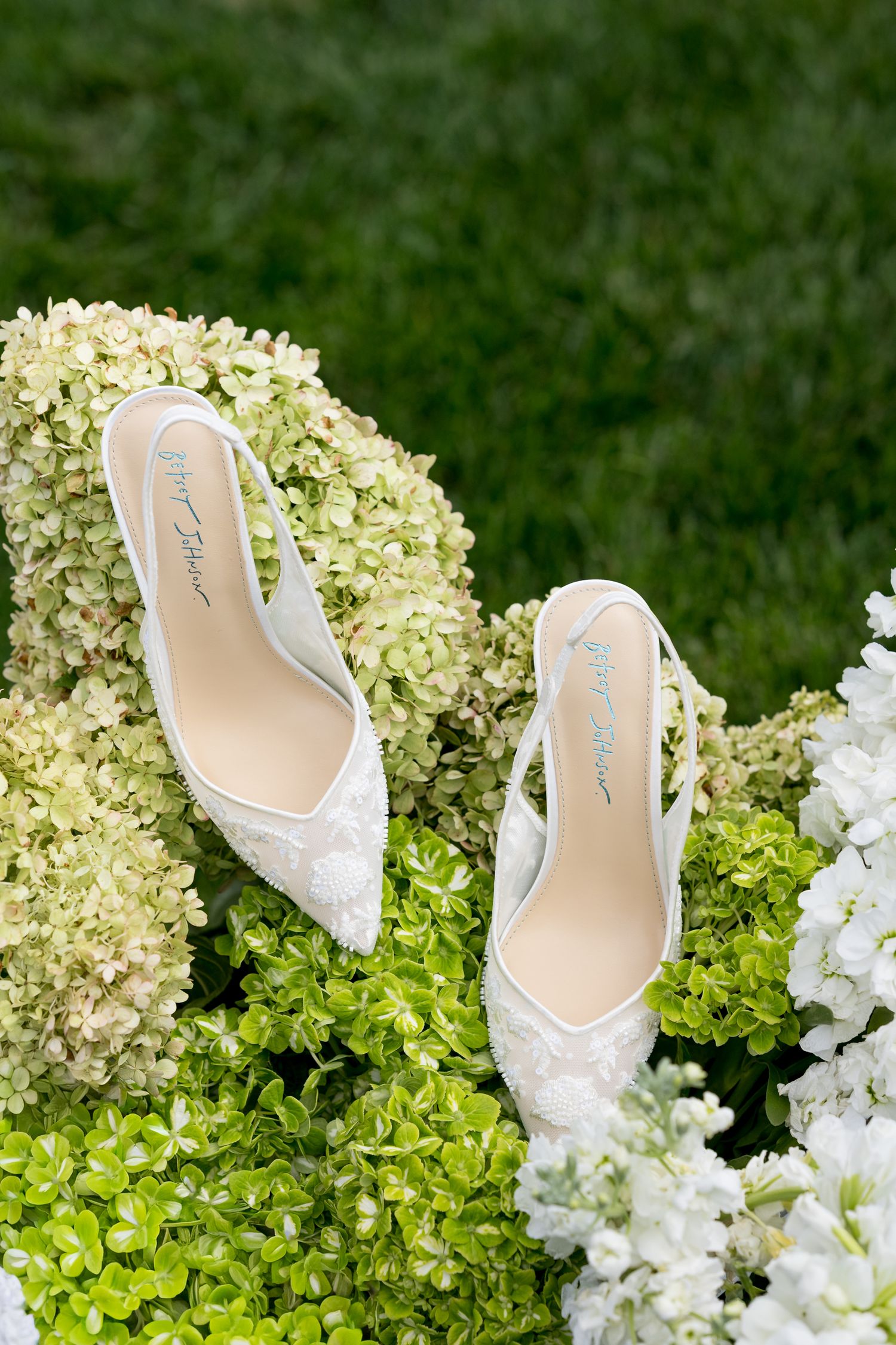 White lace bridal shoes displayed among fresh hydrangea blooms on grass.