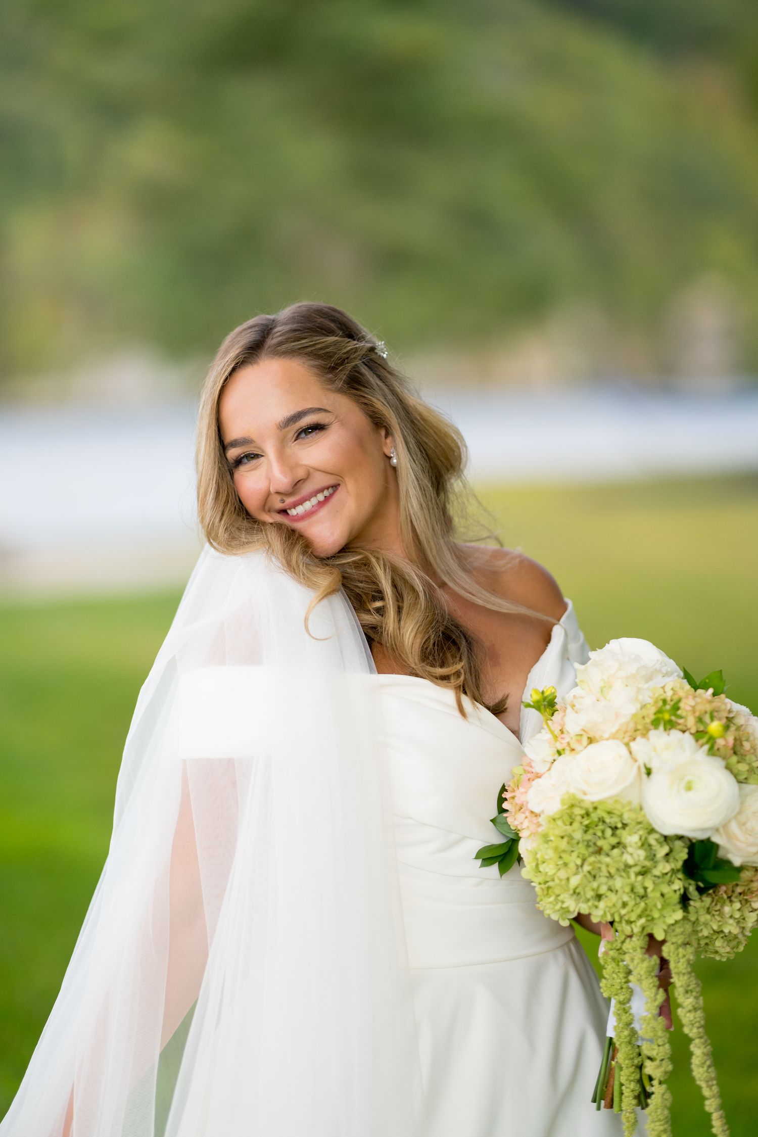 Bride in white wedding dress smiling with green hydrangea bouquet outdoors on sunny day.