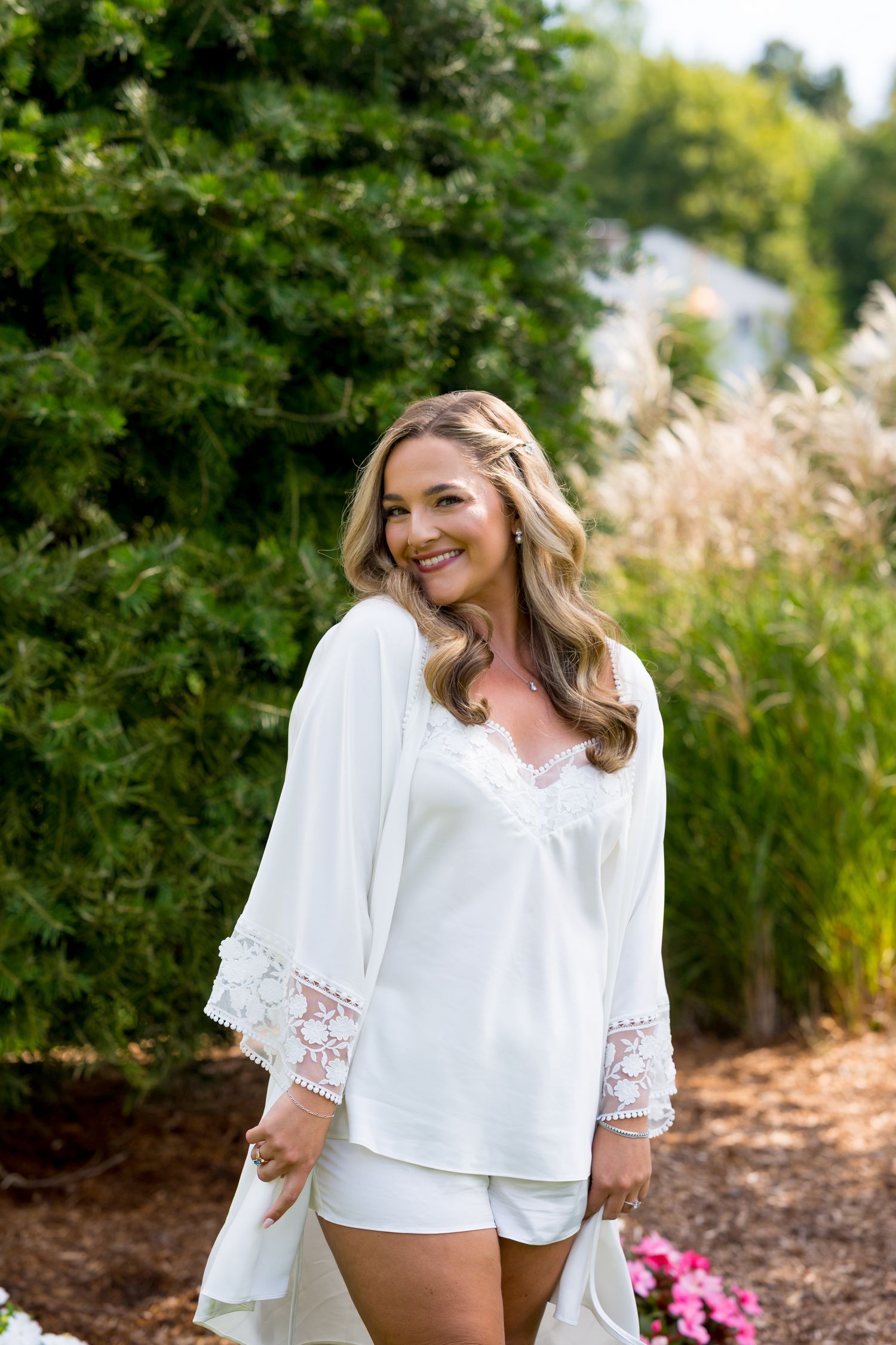 Woman in white lace-trimmed top posing outdoors against natural greenery.