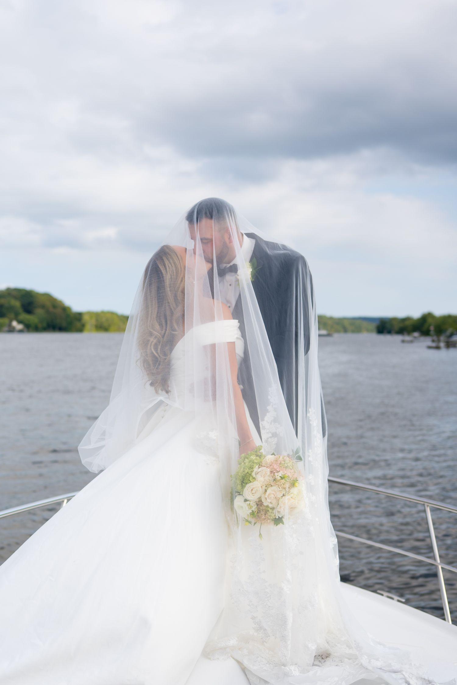 Romantic boat wedding photo with couple under veil against cloudy lake backdrop.