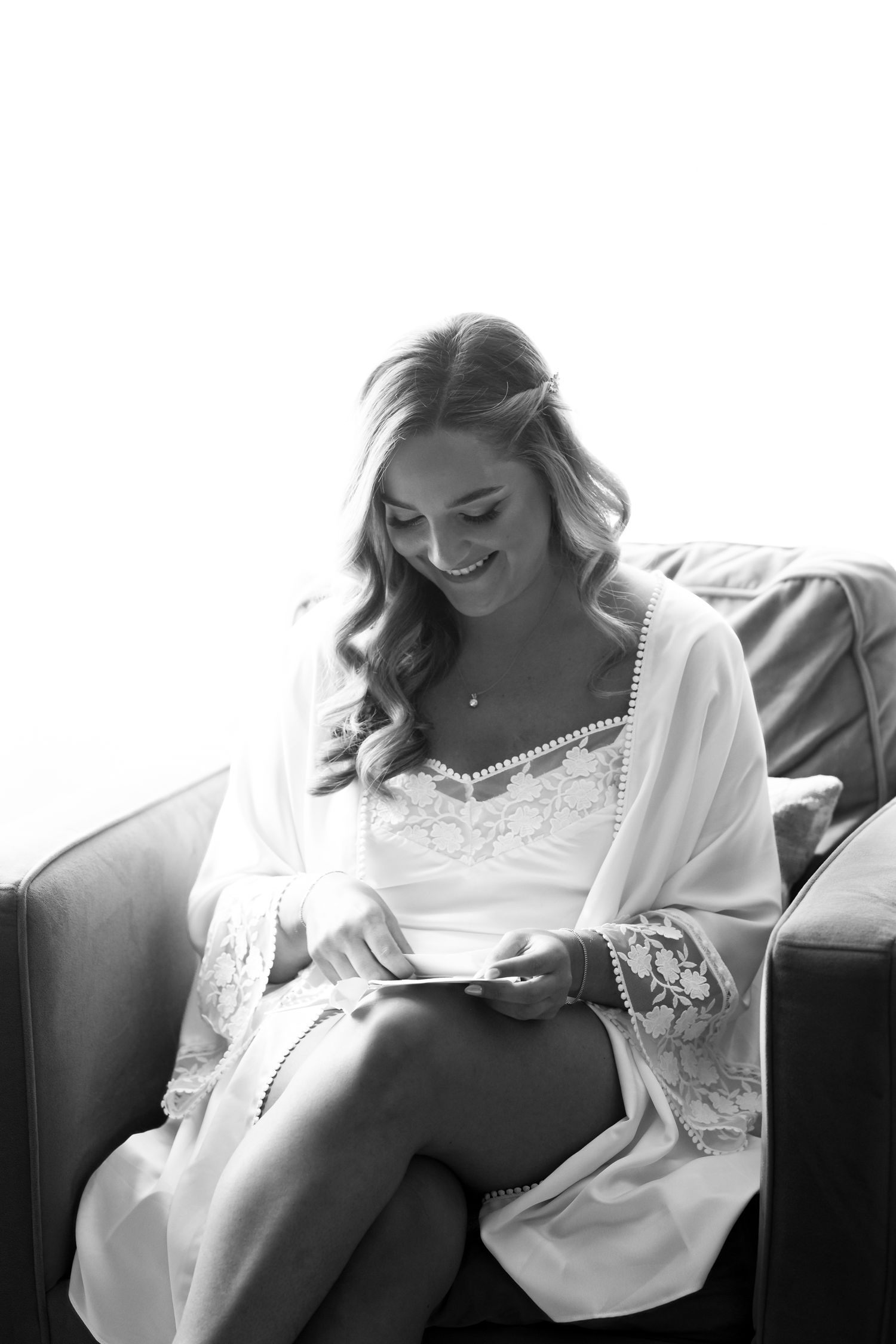 Black and white photo of someone in a white lace robe smiling while reading on a leather couch.