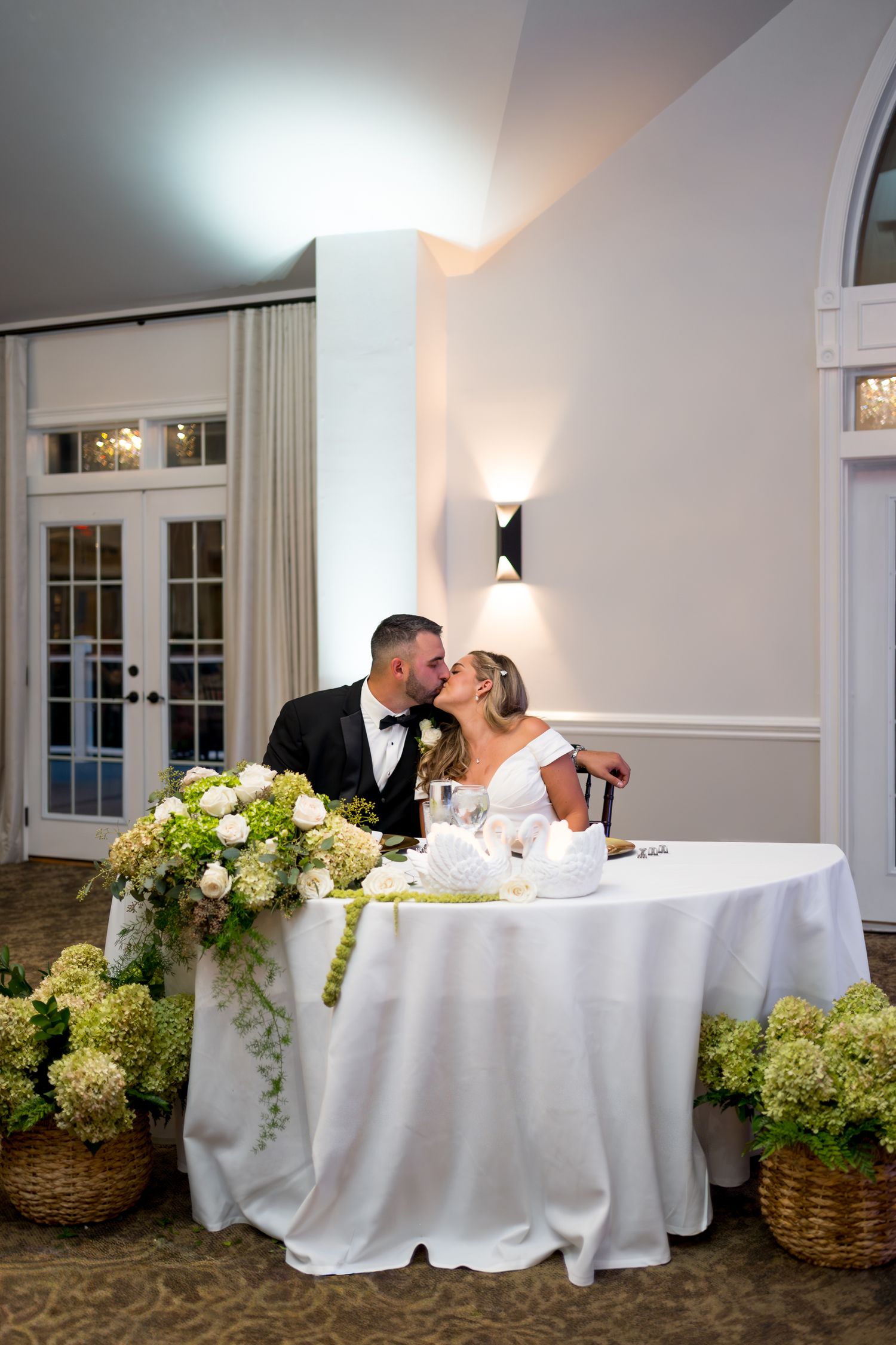 A couple shares an intimate moment at an elegant white-clothed table surrounded by lush green hydrangea arrangements.