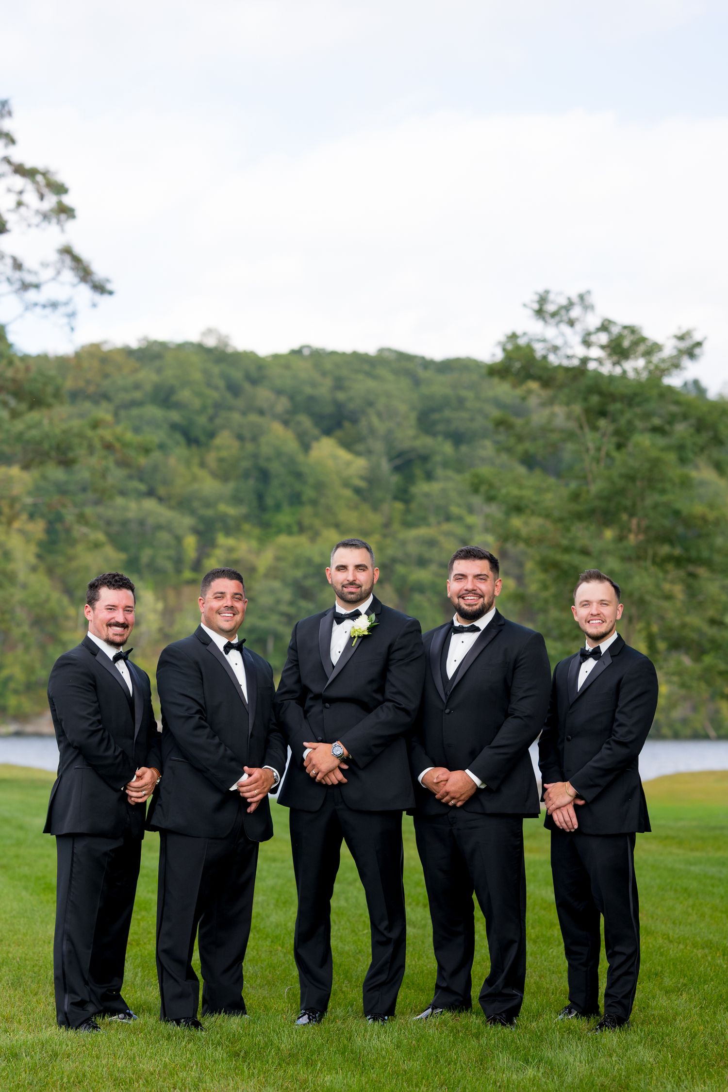A group of groomsmen in black tuxedos pose together on a grassy hill with trees in the background.