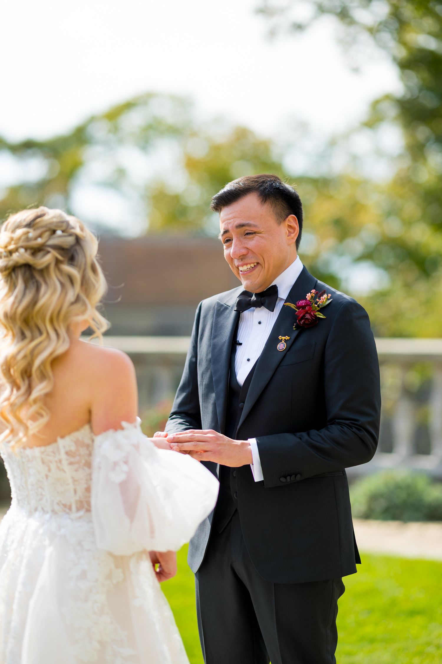 A couple shares a joyful first look moment in their wedding attire on a sunny day with green grass and trees.
