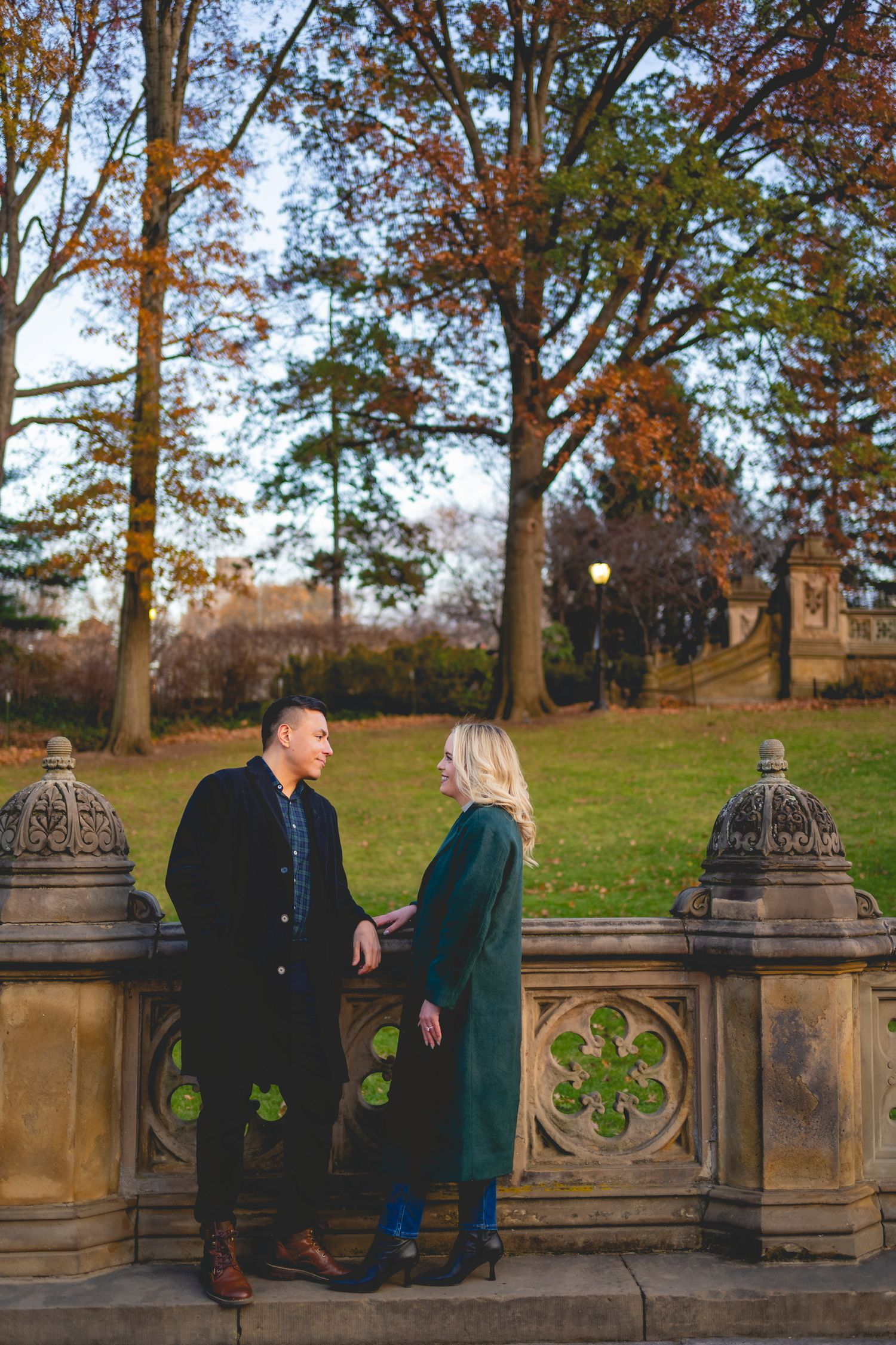 A couple stands together on an ornate stone wall during autumn with trees in the background in Central Park.