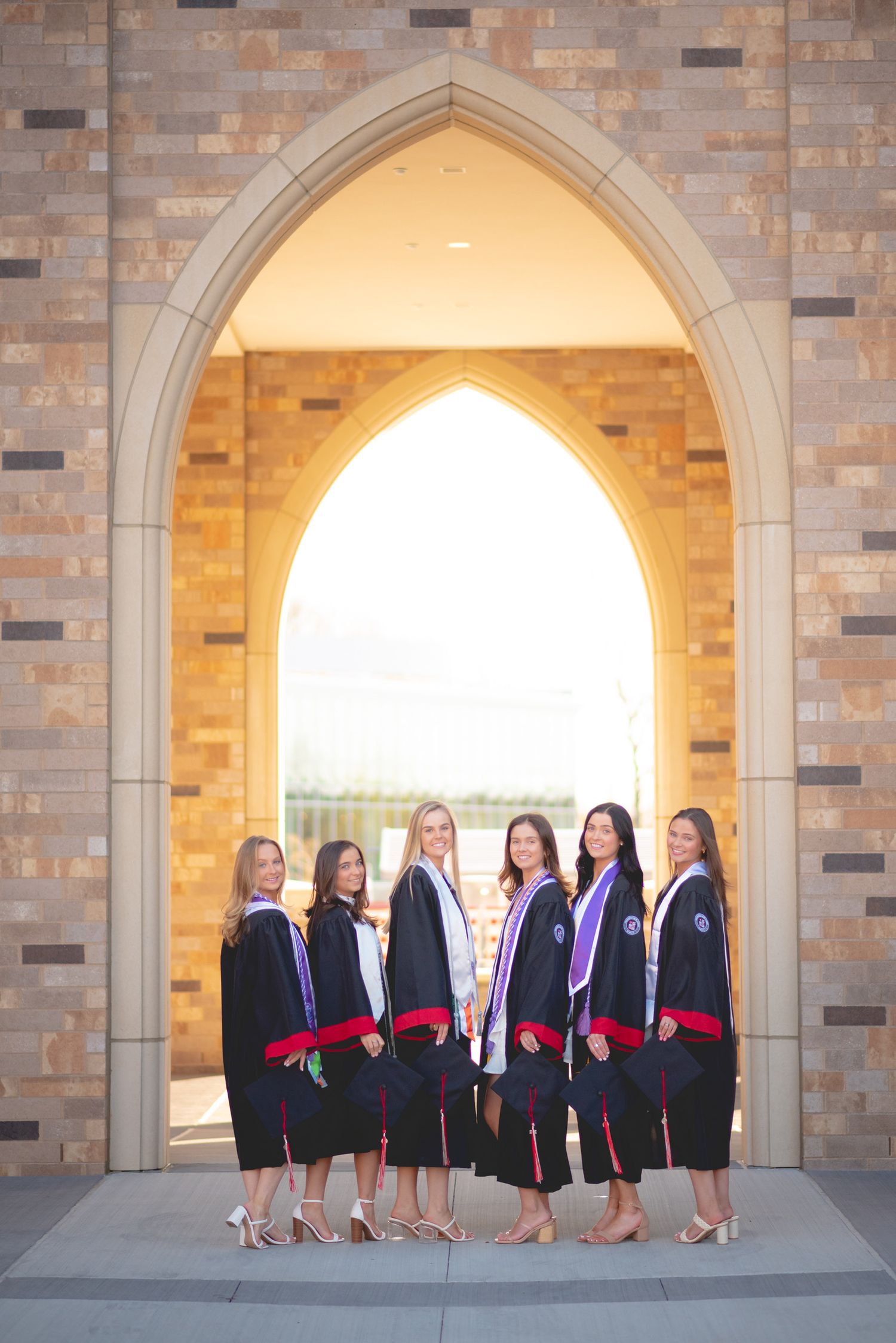 Sacred Heart University graduates in black robes pose under stone archway for graduation photos.