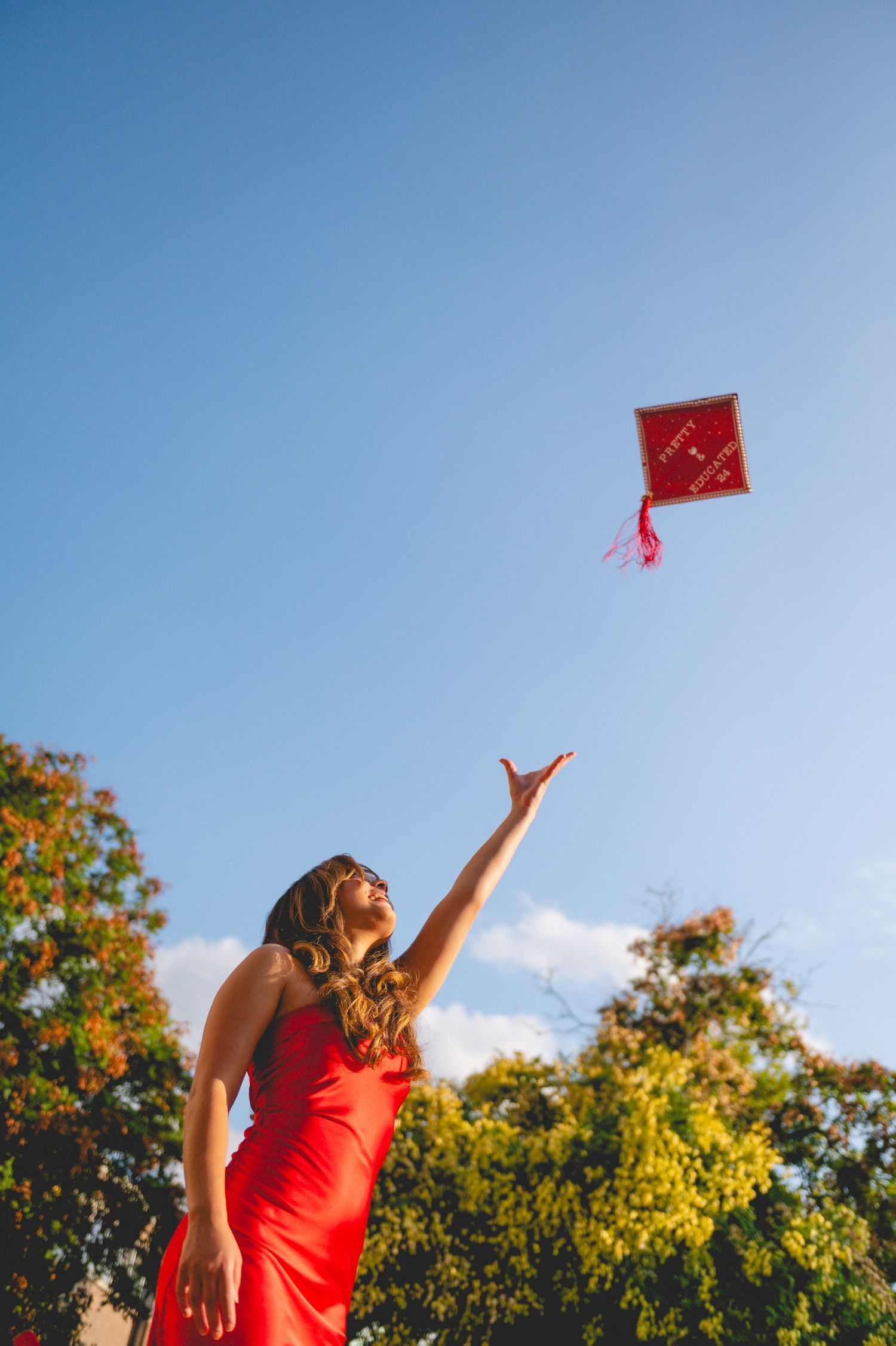 Person in red dress tosses graduation cap against blue sky and trees.