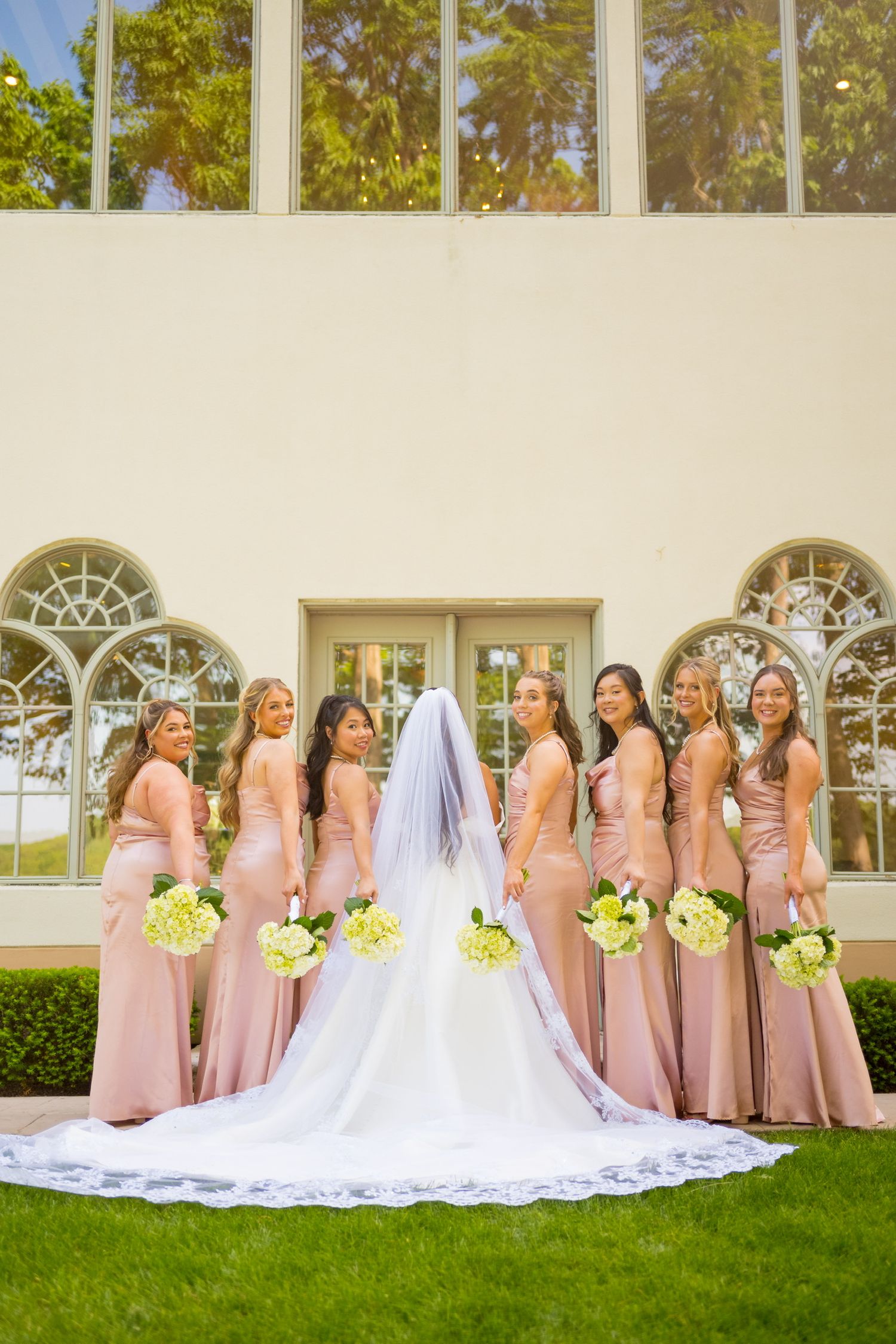 Bridesmaids in blush pink dresses pose with bride wearing flowing cathedral veil on grass.