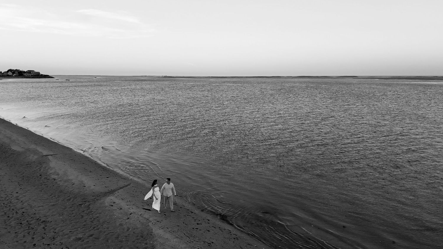 A panoramic black and white seascape shows two silhouettes walking along the shoreline at sunset.