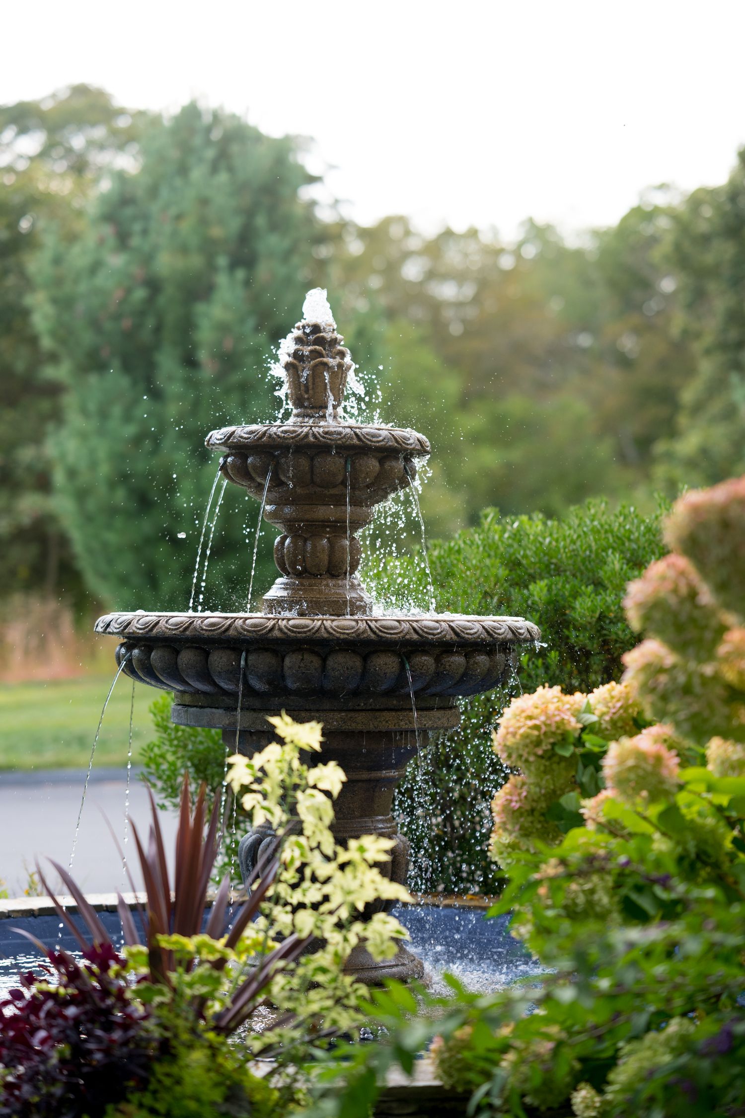 Ornate tiered fountain with flowing water surrounded by lush garden landscaping.