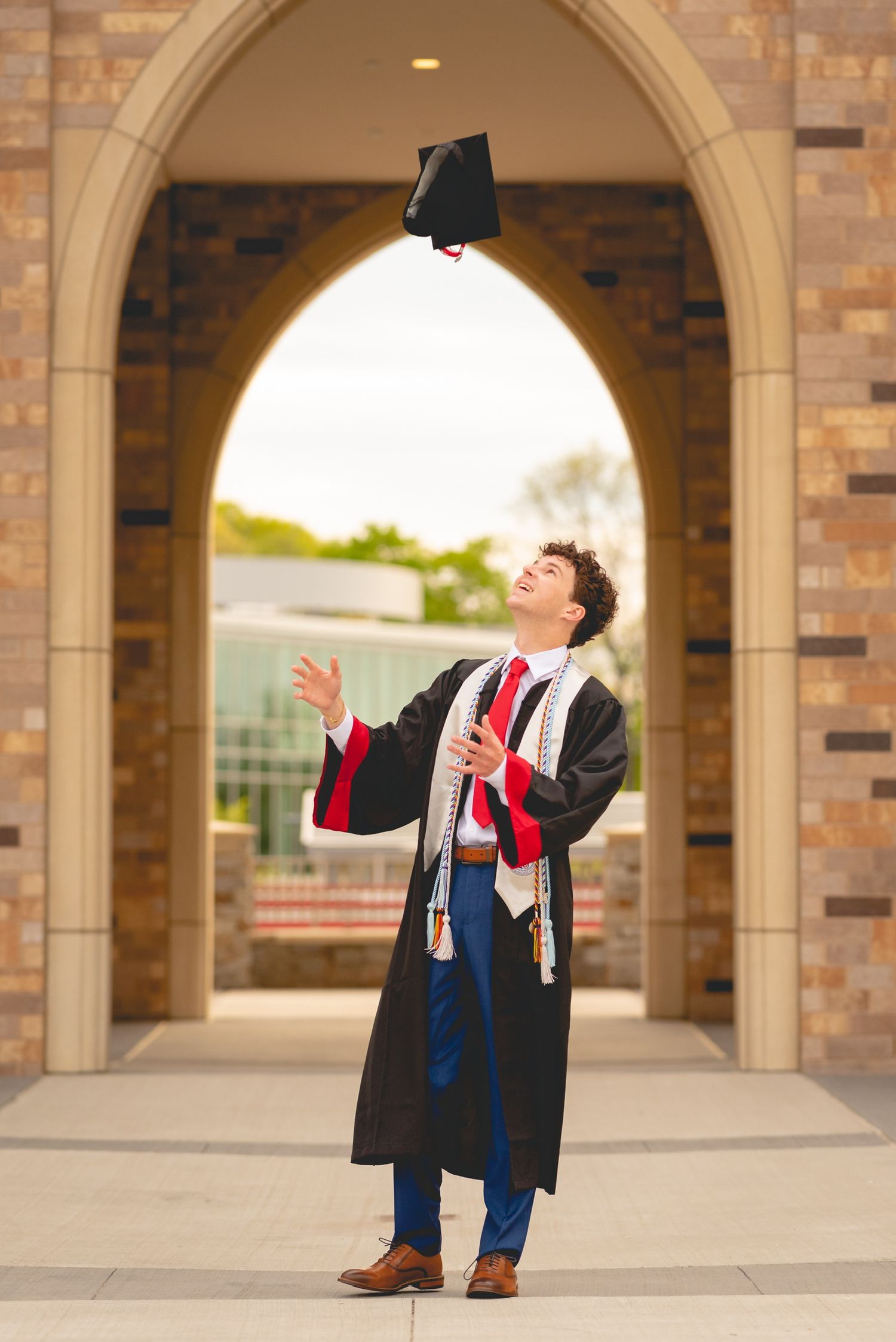 Sacred Heart University graduate in cap and gown poses joyfully under brick archways during sunset photo session.