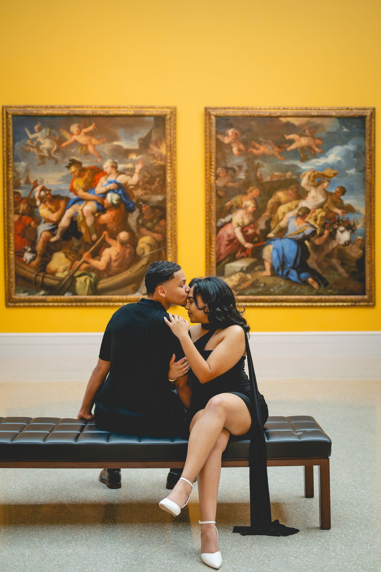 Romantic couple poses on art gallery bench with yellow walls and classical artwork in background.