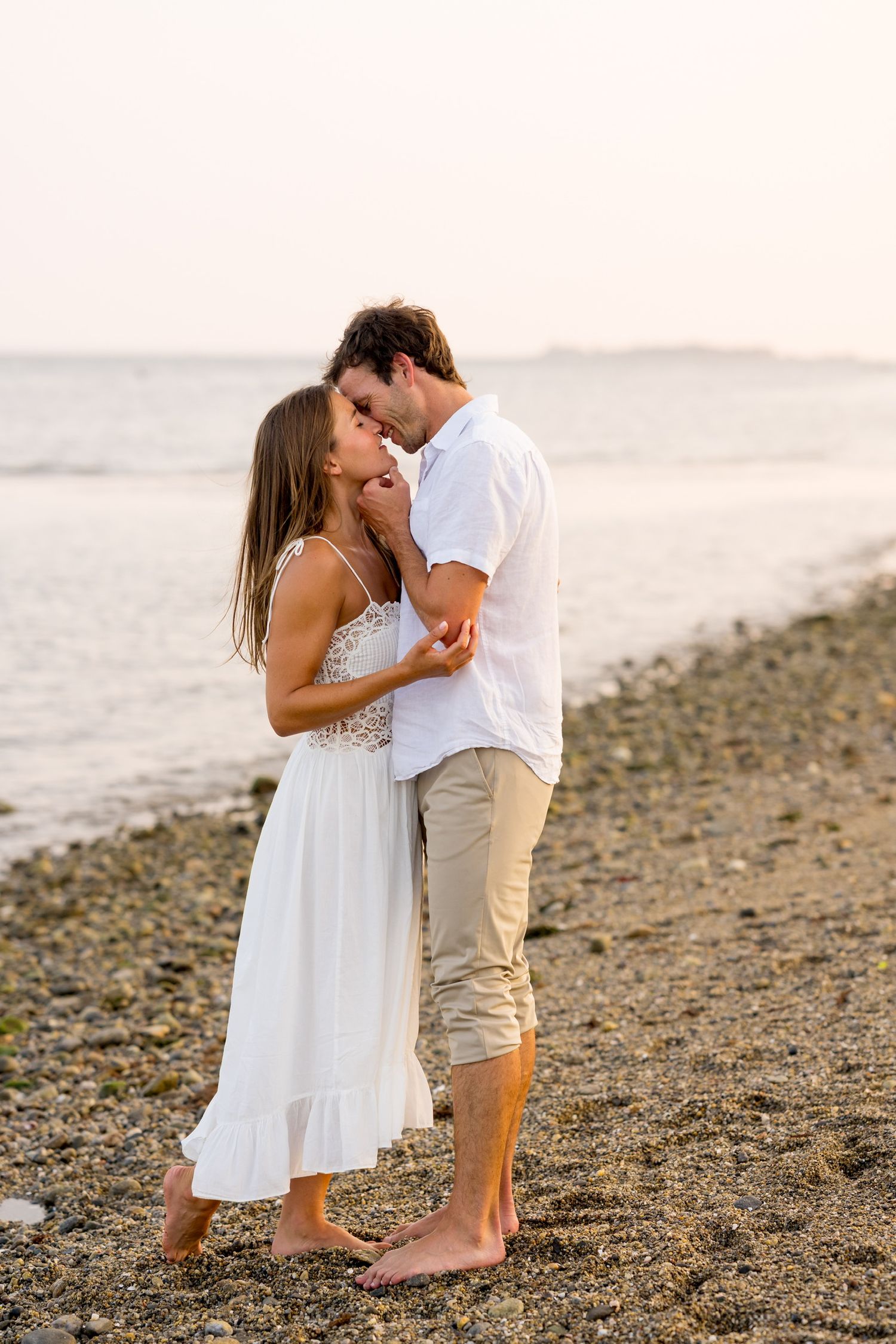 Romantic couple in white outfits sharing intimate moment on rocky beach at sunset in Westport, Connecticut.