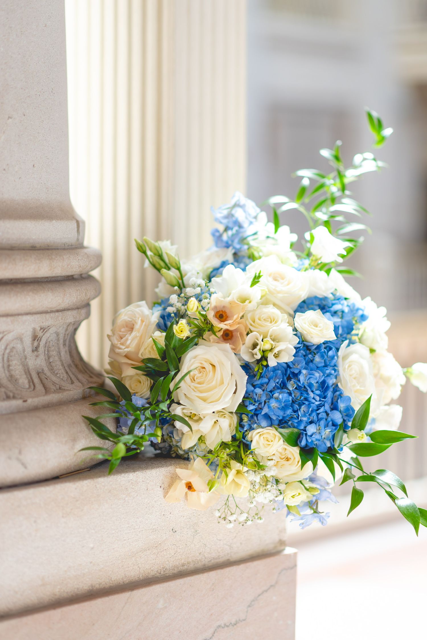 Blue hydrangeas and cream roses arranged on stone ledge with classical columns.