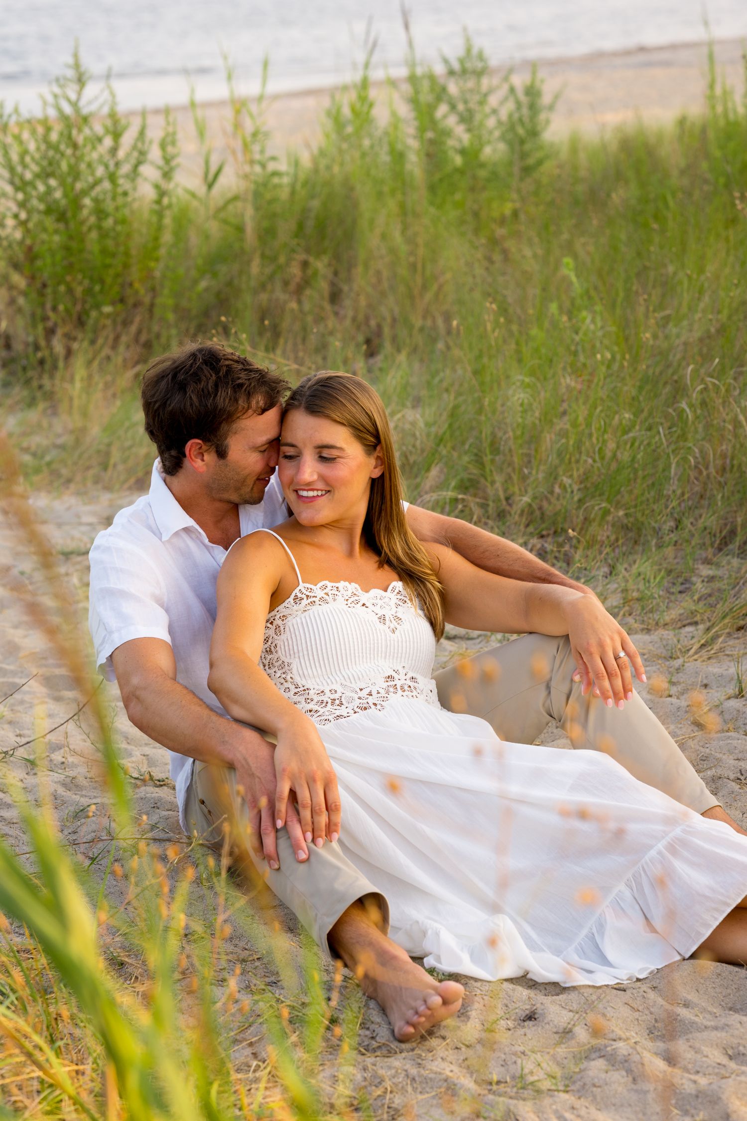 A romantic couple embraces on a sandy beach with tall grass in the background during golden hour.