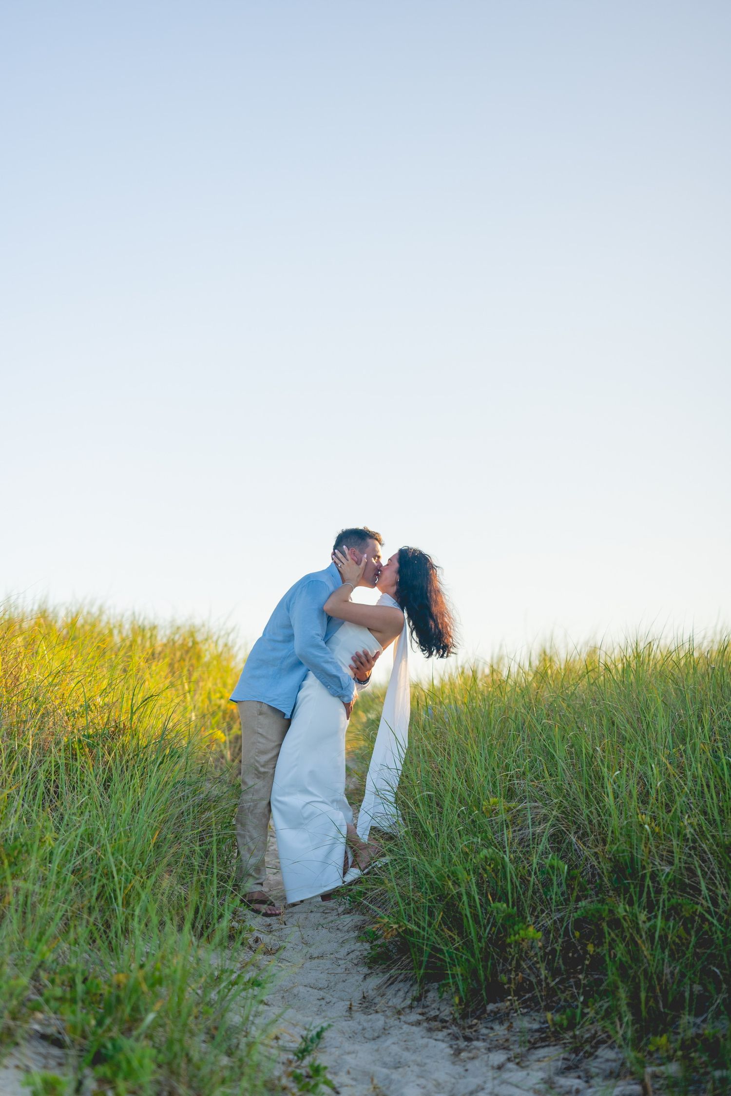 A couple shares a romantic moment among tall beach grass at sunset at Chatham Lighthouse Beach in Chatham, Massachusetts.