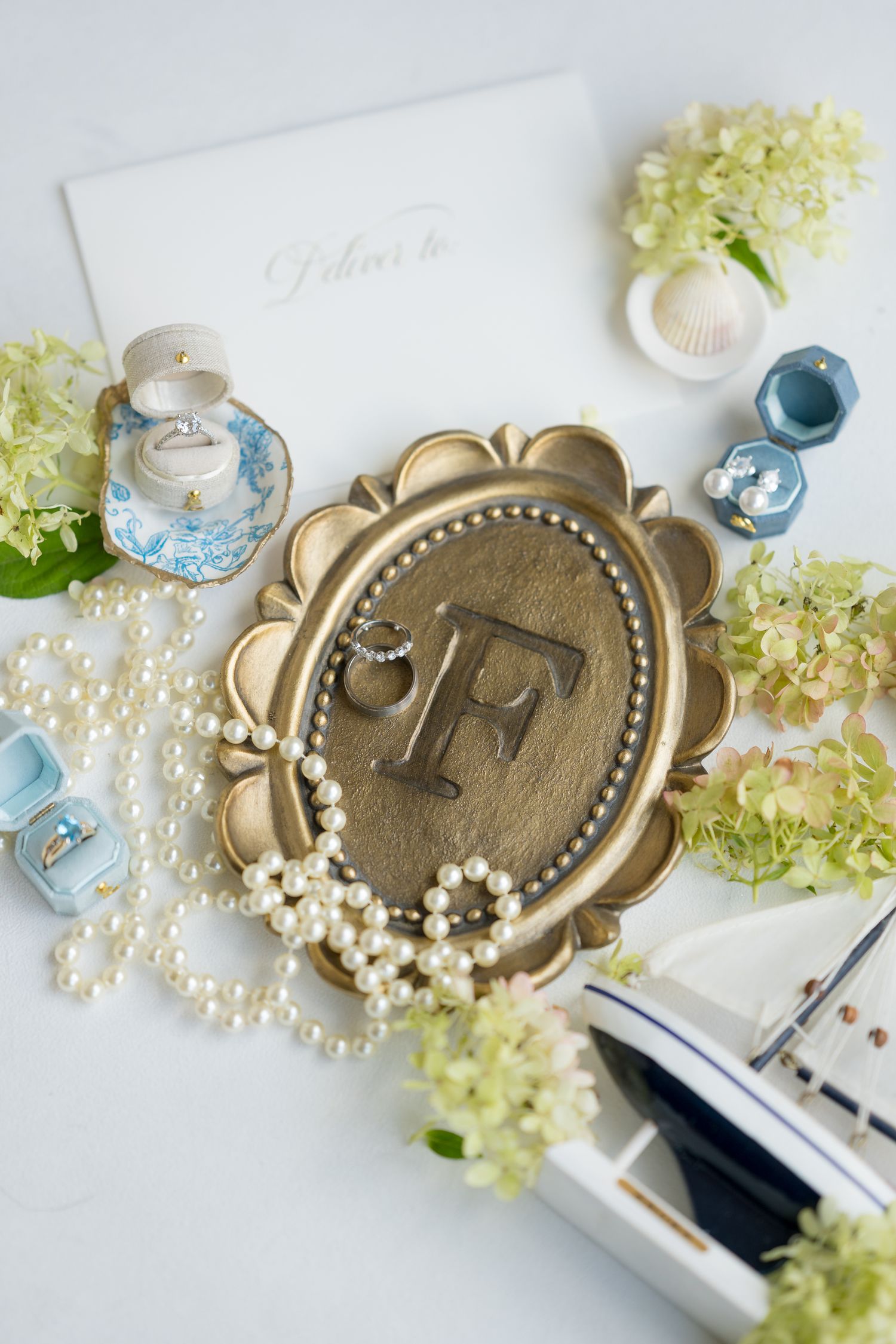 Wedding details flatlay with monogram medallion, pearls, and hydrangeas in soft blue and white.