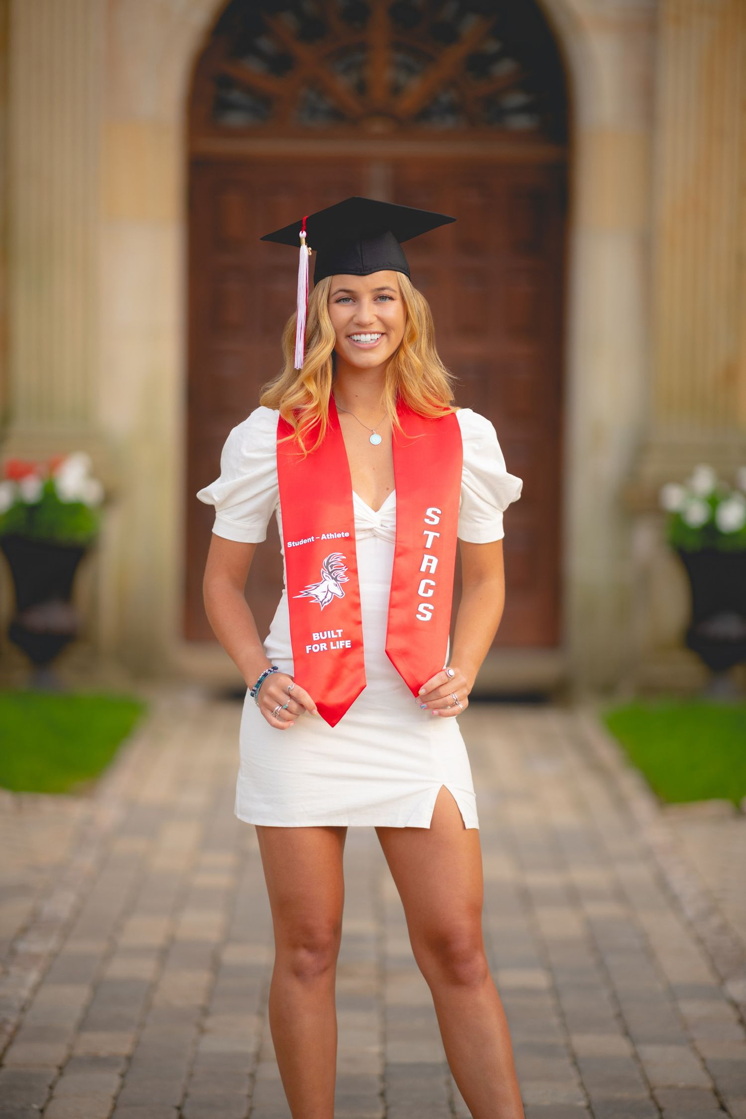Graduate in white dress and red stole poses for celebratory photos on brick pathway near ornate building.