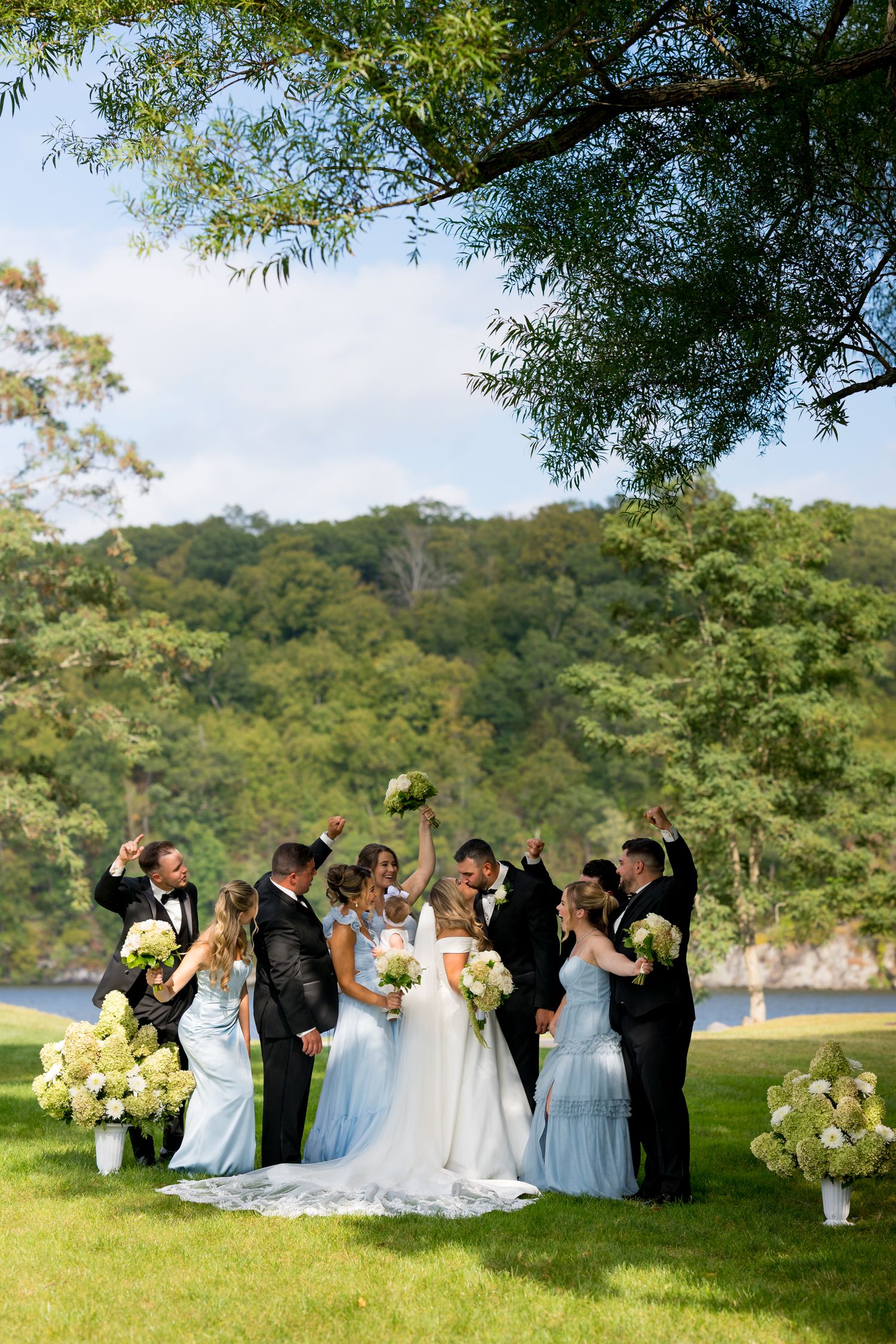 Wedding party gathered on a grassy lawn with trees and water in the background.